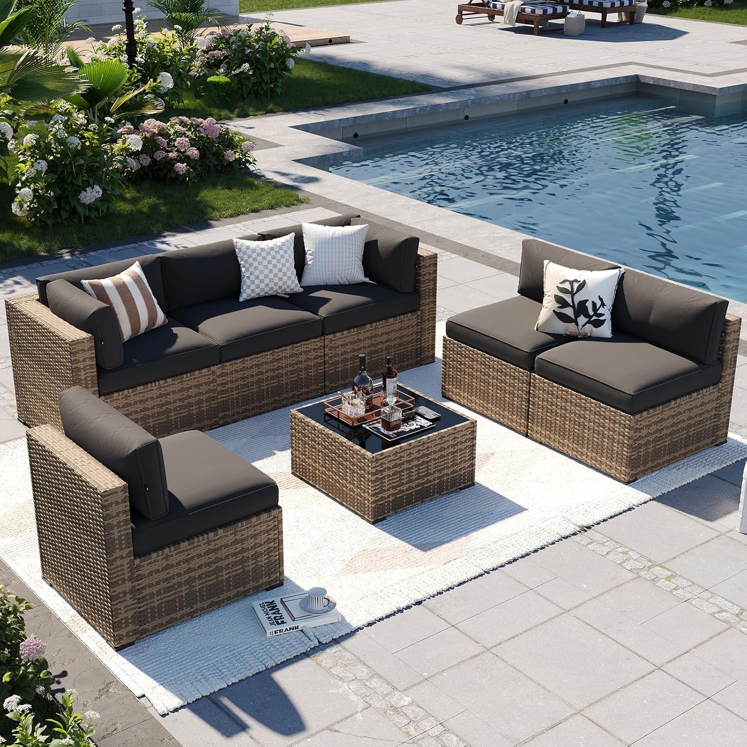 Outdoor patio area with wicker furniture including a sofa, two chairs, a small table, and cushions on the furniture, placed on a white rug. A swimming pool with steps is visible in the background. There are plants and flowers along the edge of the patio.