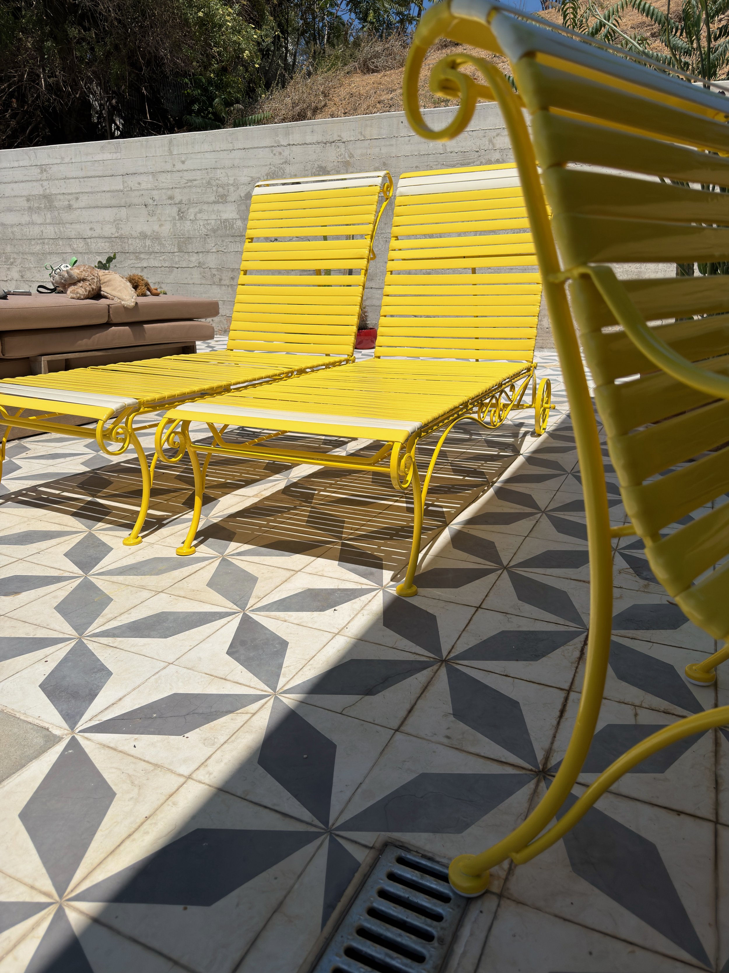 Yellow metal lounge chairs on a patterned tile floor with a concrete wall in the background. There is a brown outdoor sofa with plush toys on it to the left.