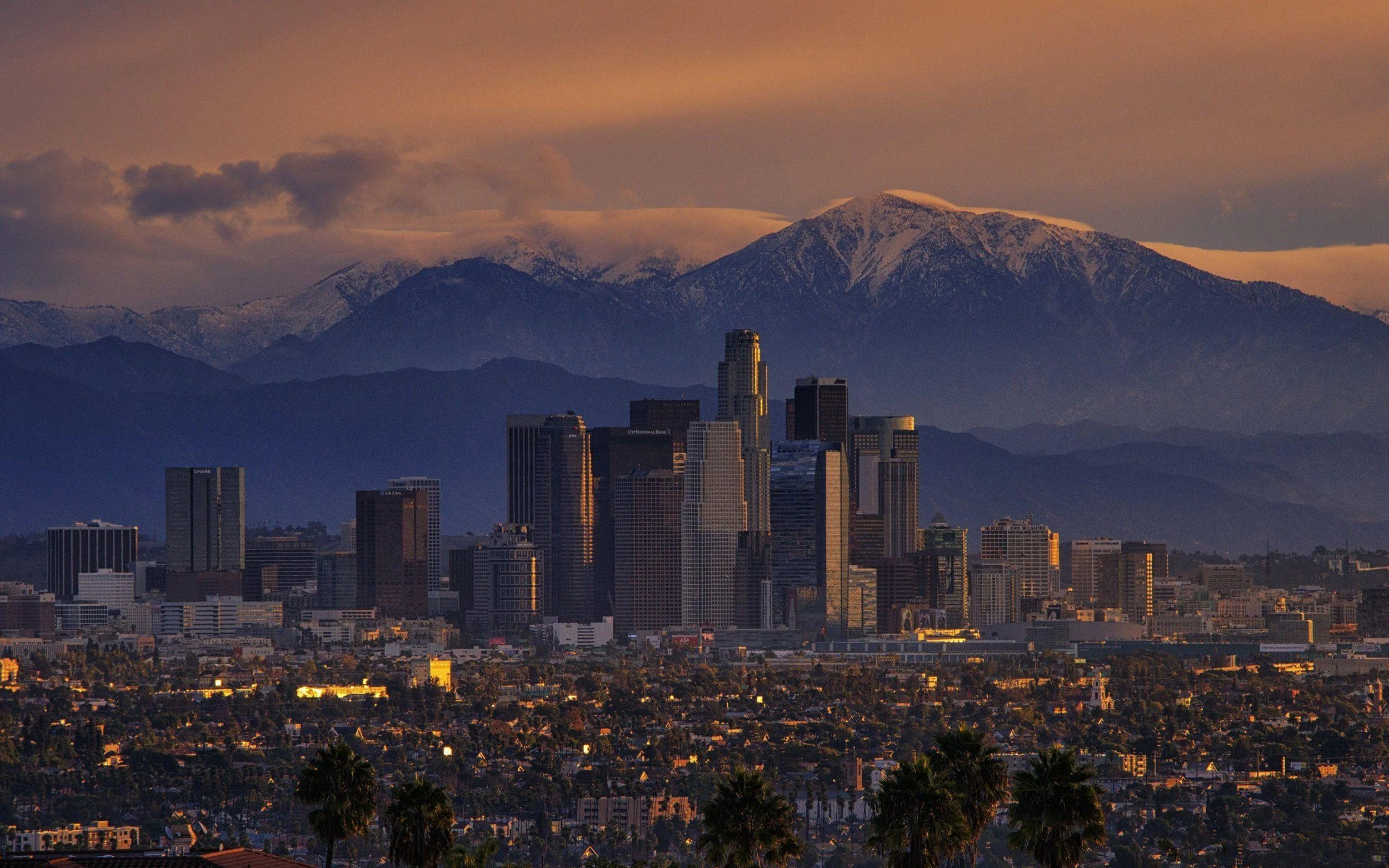 Downtown Los Angeles skyline with high-rise buildings and mountains in the background, under a pinkish-orange sky at sunset or sunrise.
