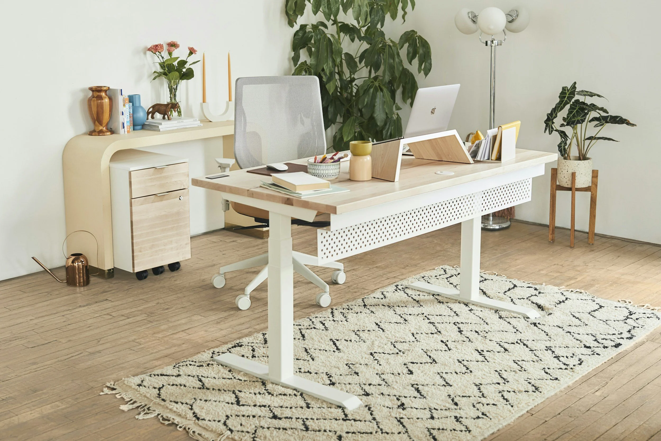 A modern office space with a white desk, gray office chair, and decorative elements including potted plants, books, a basket with pens, and a white lamp. The floor is wooden, and there's a black and white patterned rug under the desk.