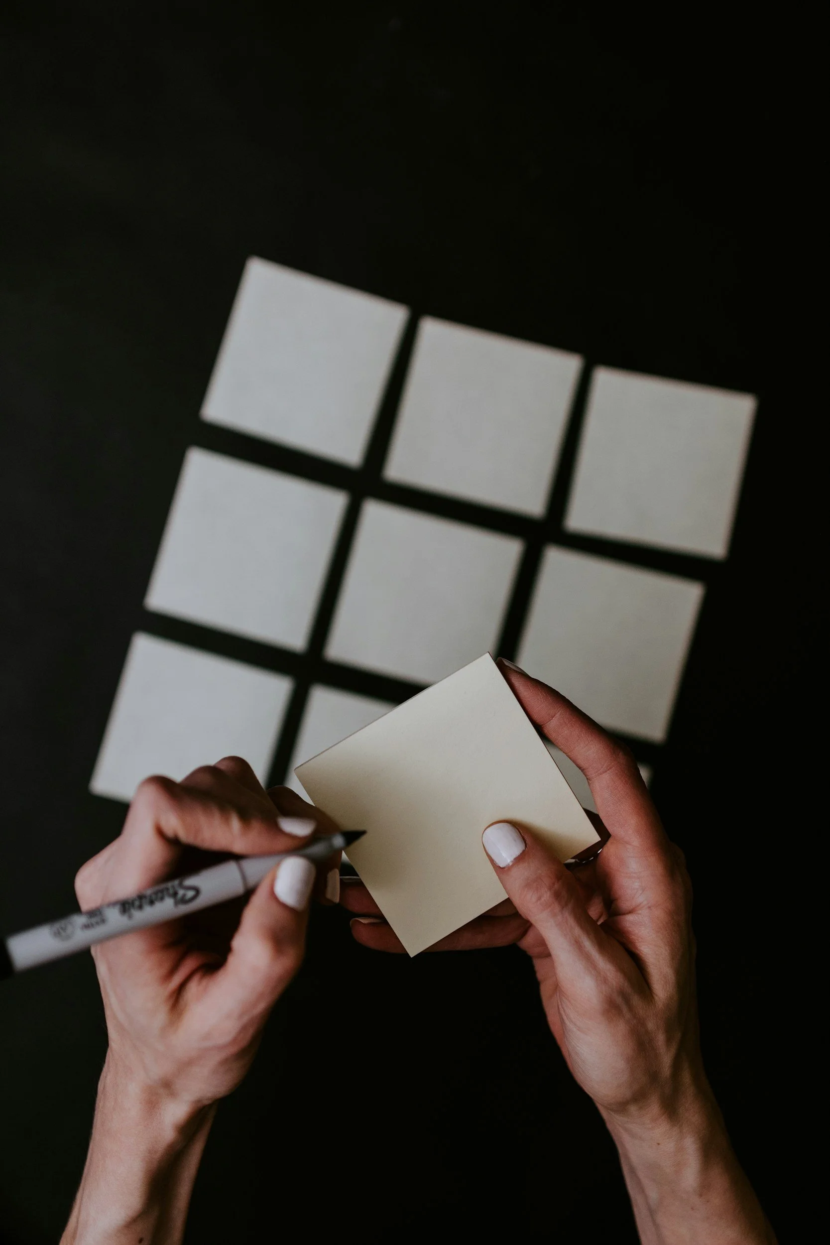 Person drawing on a small notepad with a black marker, with a black background and partially visible white square stickers on the surface.