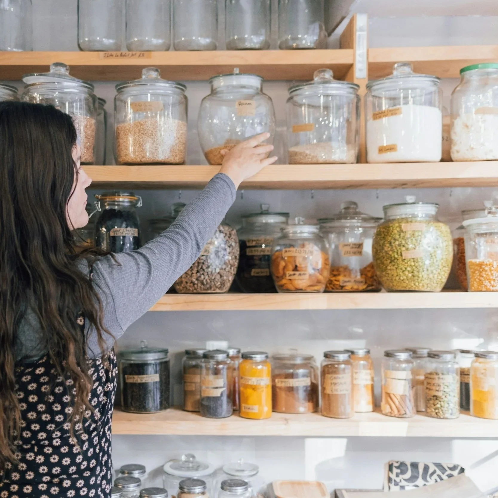 A woman with long dark hair wearing a gray sweater is reaching for a glass jar filled with an orange substance on a wooden shelf. The shelf holds multiple glass jars and containers filled with various dry goods, such as grains, seeds, and spices, in a pantry or storage room.