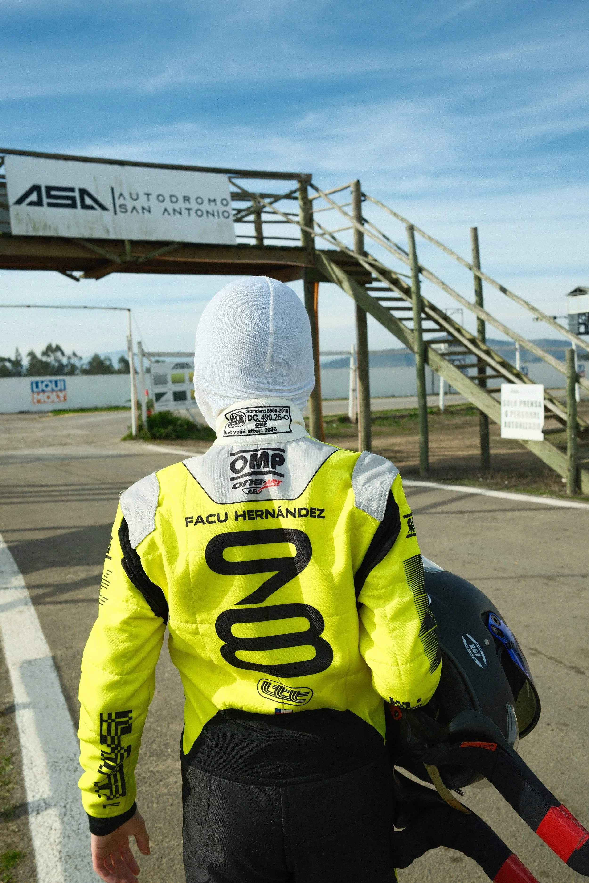 Persona de carrera en pista de autos con chaqueta amarilla y casco en mano, de espaldas, en el Autódromo San Antonio.