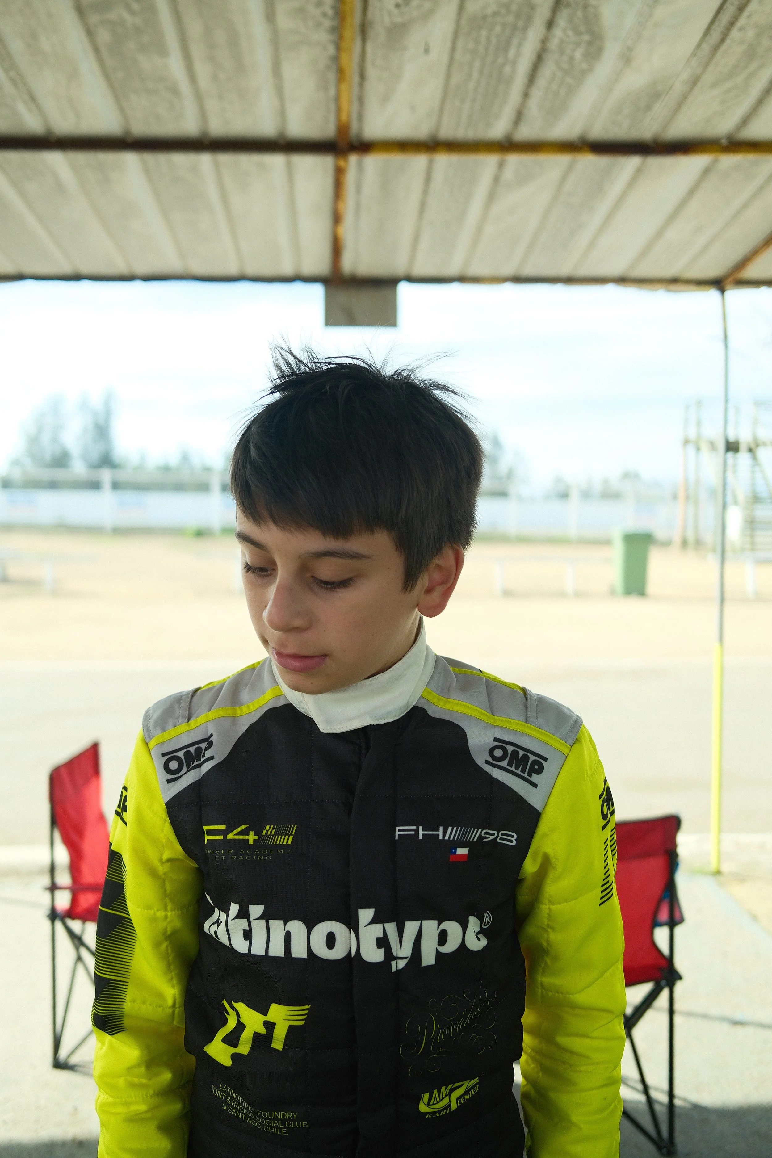 Joven piloto con uniforme de carreras, en un entorno al aire libre con sillas de camping y fondo borroso.