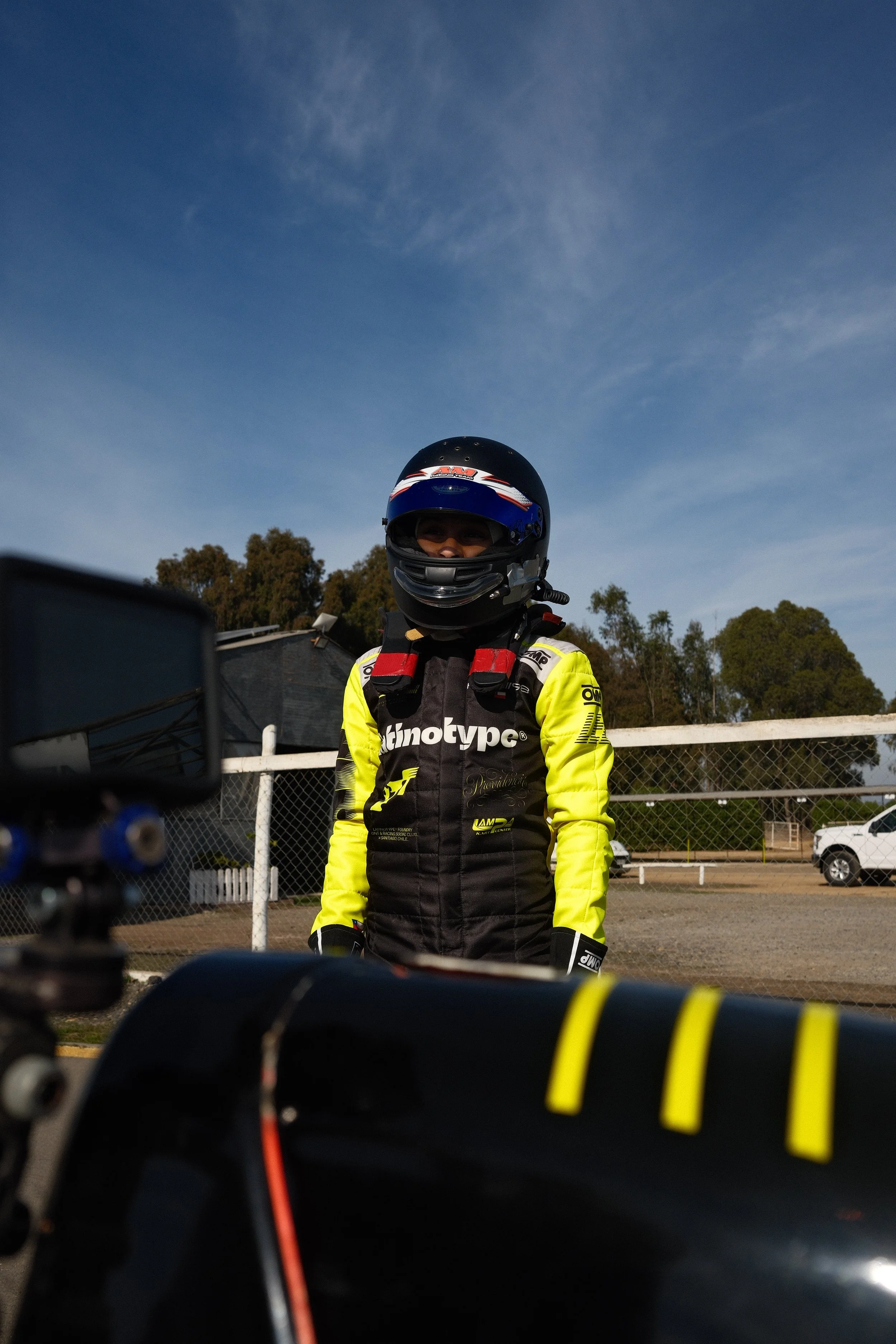 Persona con casco y traje de carreras en una pista de auto, con fondo de cielo y árboles.