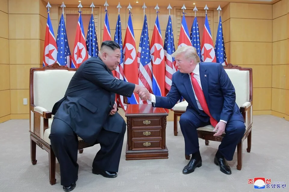 Democratic People's Republic of Korea President Kim Jong Un shakes Donald Trump's hand with North Korean and American flags arrays side by side in the background.