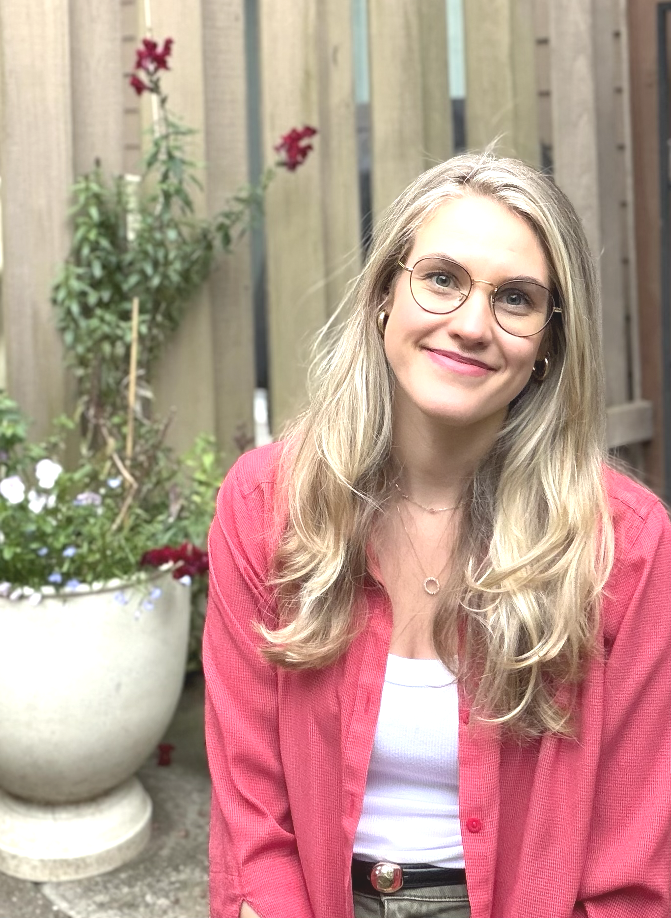 A woman with long blond hair wearing glasses and a pink jacket, smiling, in front of a wooden fence and potted plants with red and white flowers.