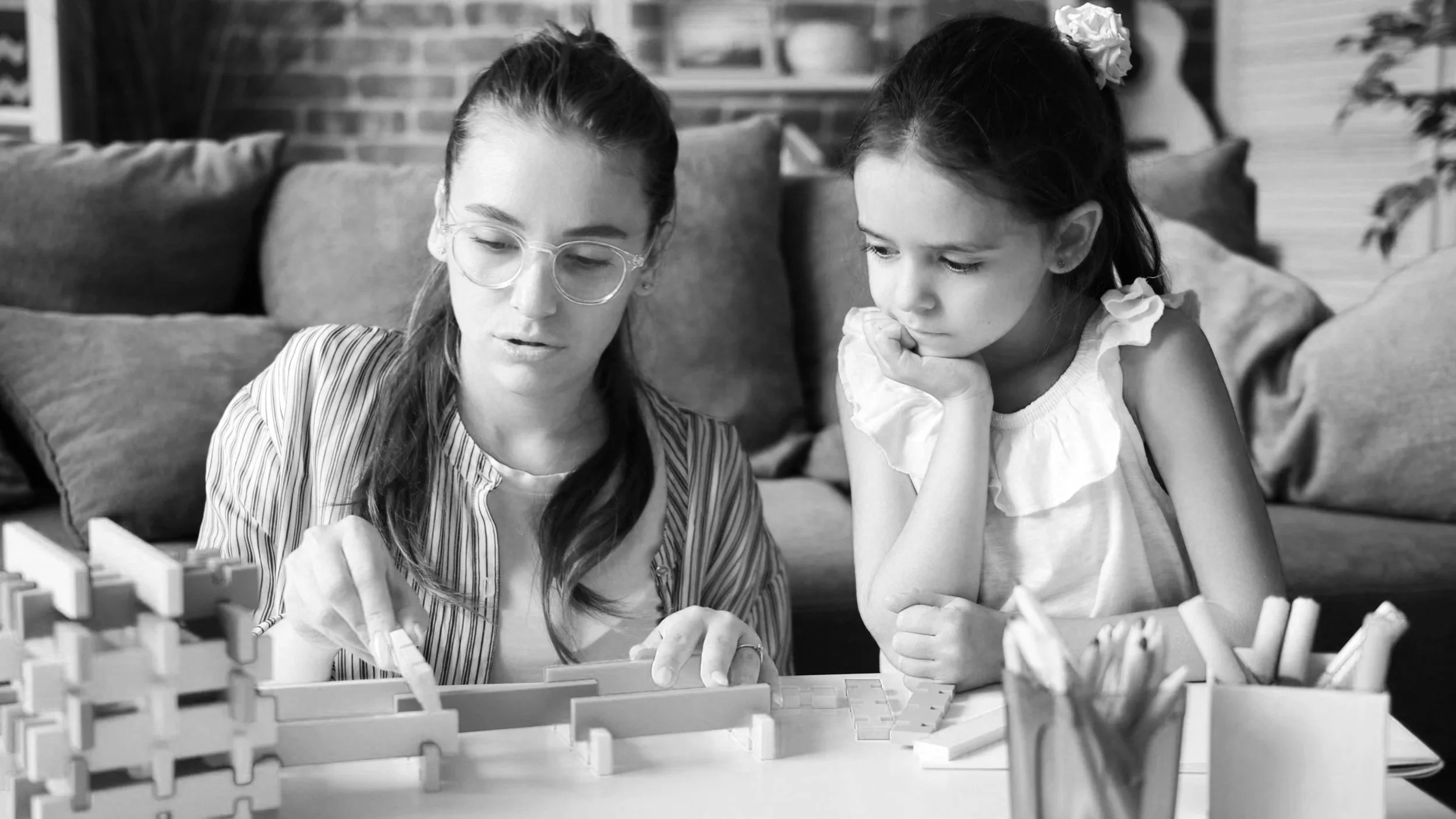 A woman with glasses and a young girl are playing with building blocks on a table in a living room. The woman is showing something to the girl, who is looking attentively. They are seated on a couch with cushions, with a brick wall and a window in the background.
