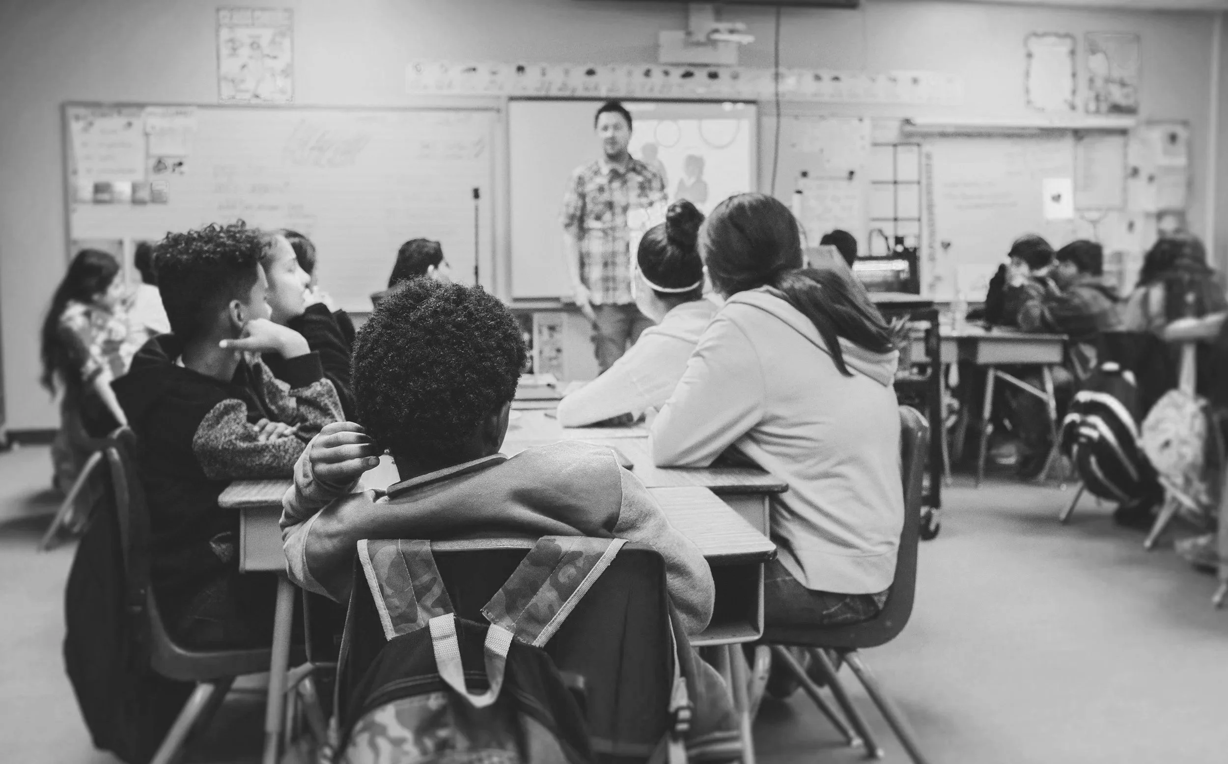 high school classroom with a male teacher standing and male and female students sitting attending to the instruction. Students represent a diverse student body; male and female, white, African American, and Hispanic