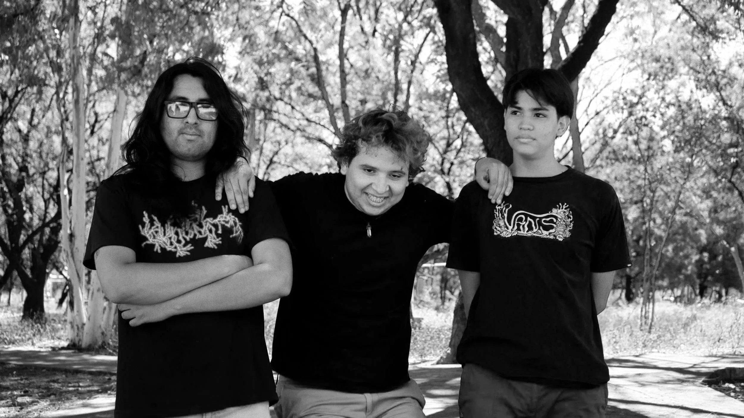 Three boys standing outdoors under trees, arm around each other's shoulders, smiling and looking relaxed