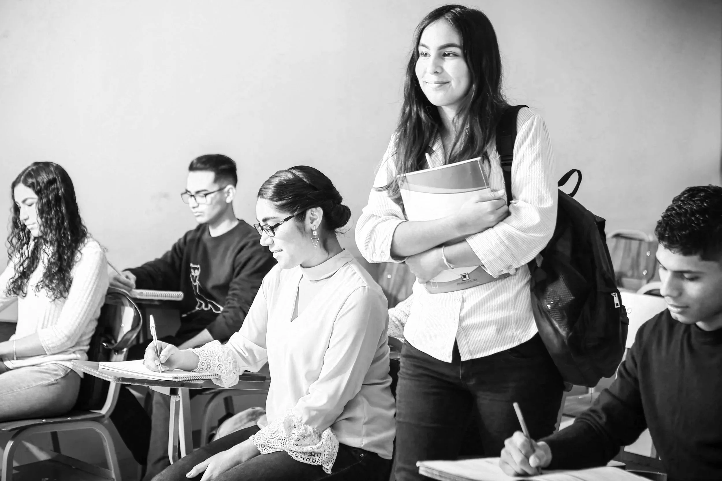 white female college student standing with book in a classroom with other co-ed students sitting at desks