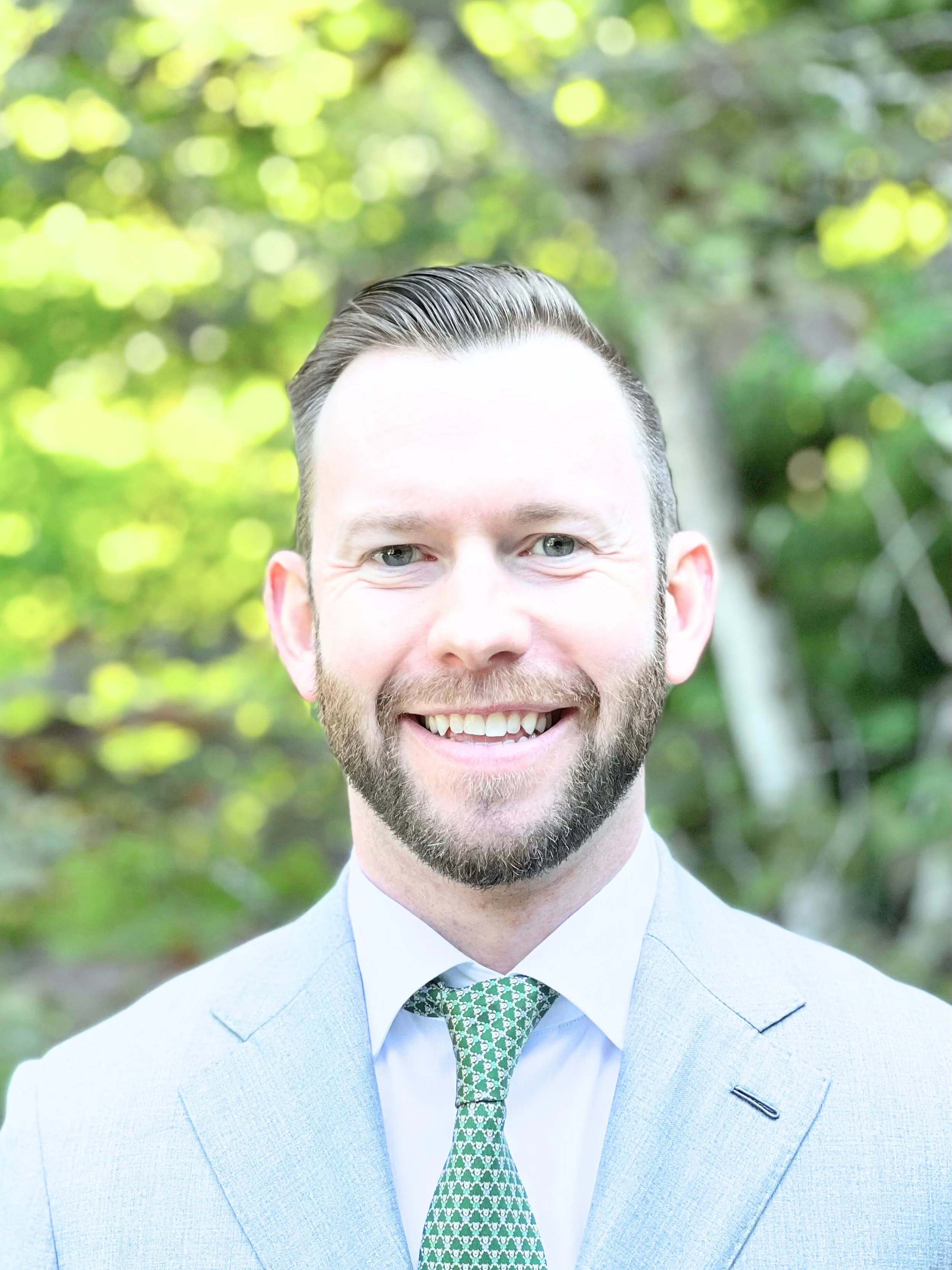A smiling man dressed in a light grey suit, white shirt, and green patterned tie outdoors with a blurred background of green and yellow foliage.