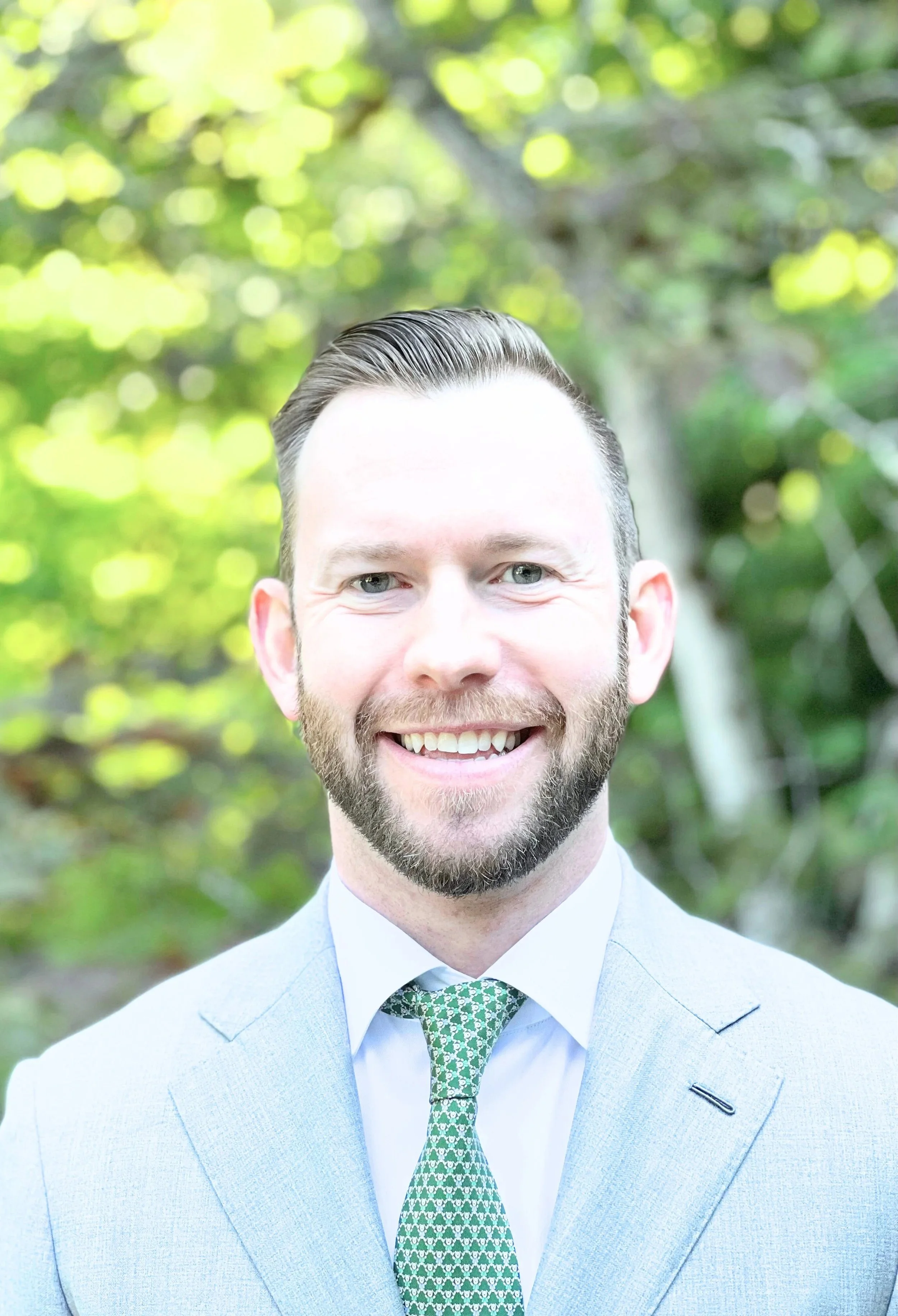 A young man with a beard and blue eyes, smiling, dressed in a light blue suit, white shirt, and green patterned tie, outdoors with a blurred green leafy background.