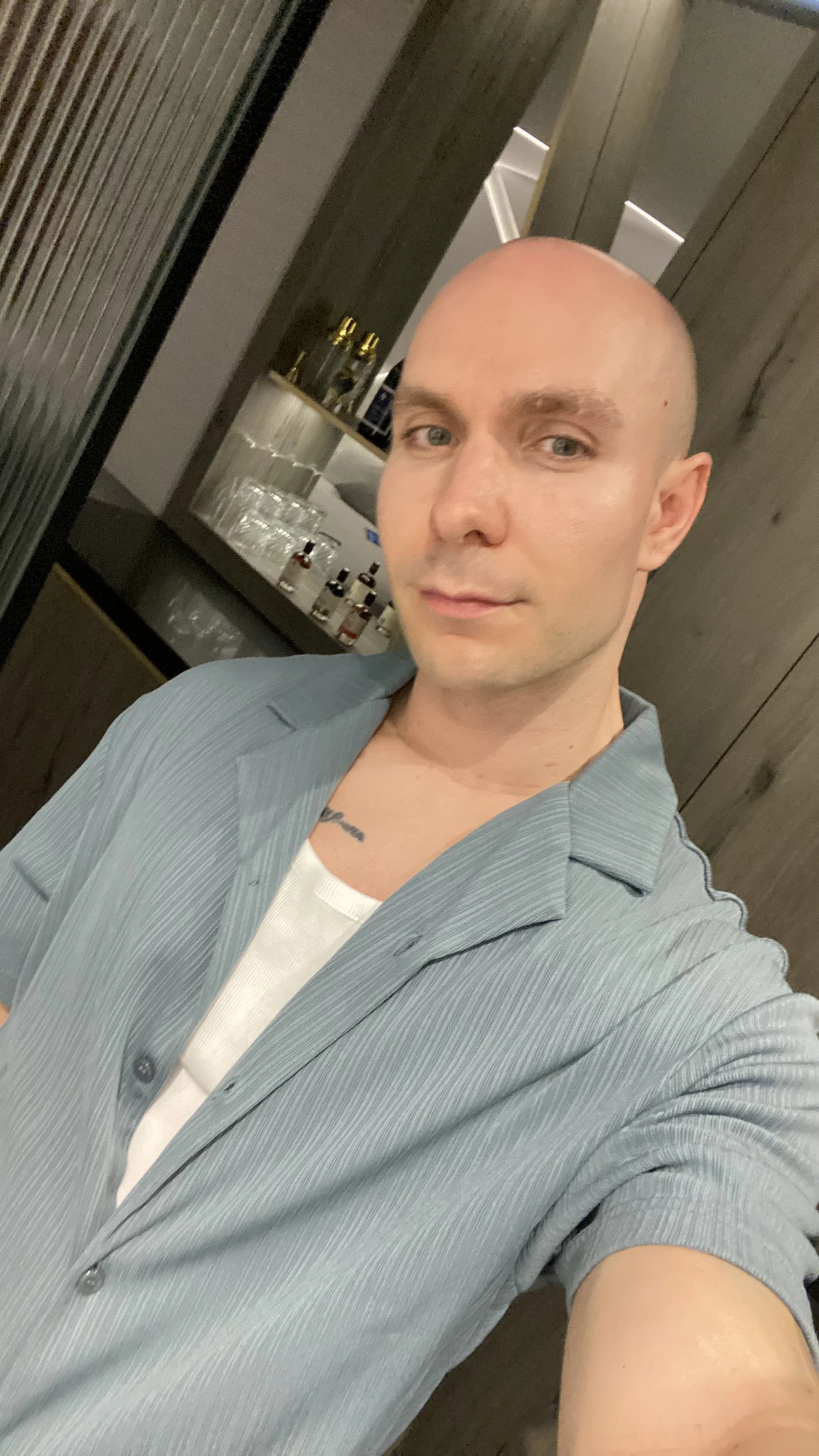 A man with a shaved head and light blue shirt taking a selfie in front of a mirror inside a room, with a wooden wall and a shelf with glasses and bottles in the background.