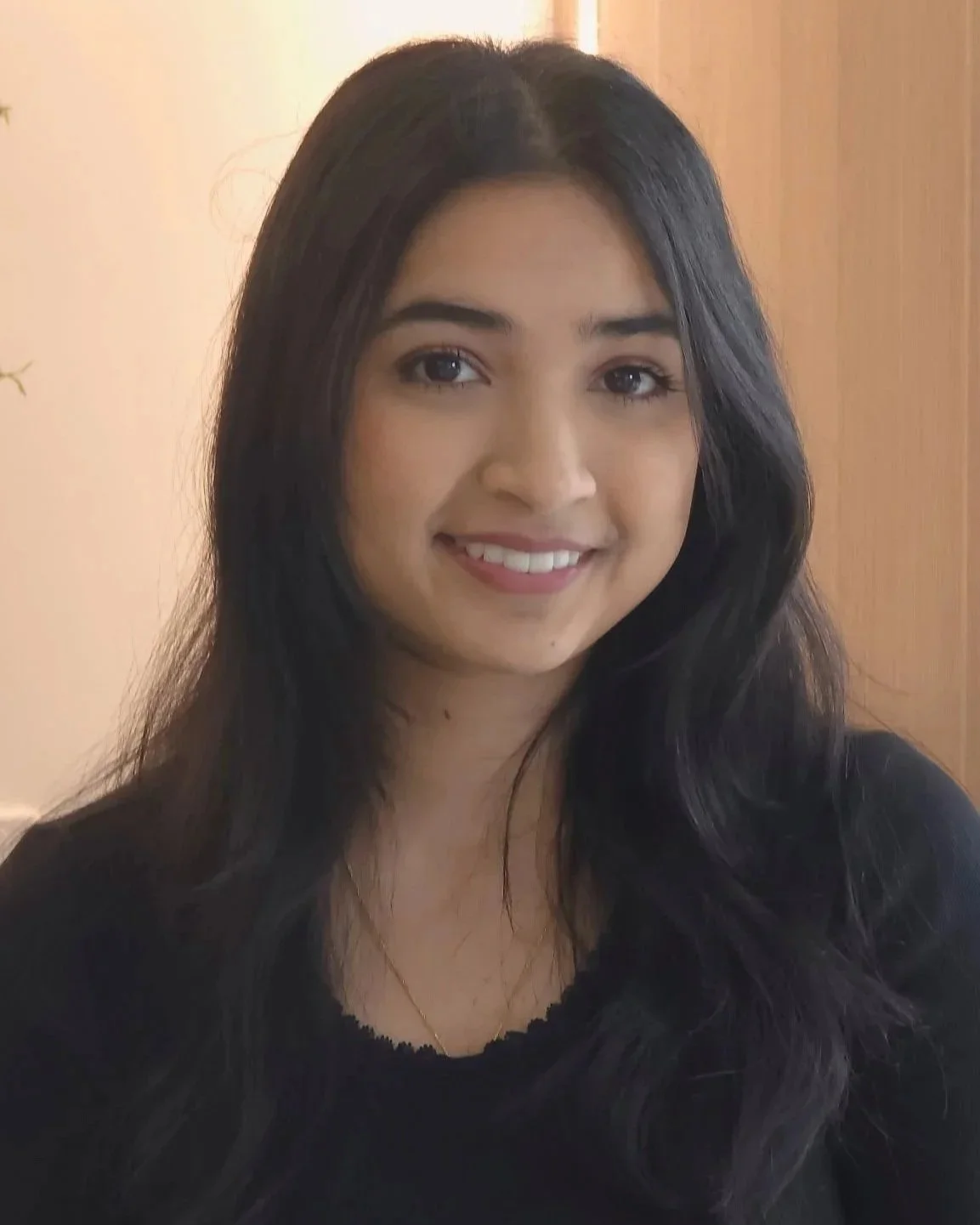 A young woman with long black hair, smiling, wearing a black top, indoor background with wood paneling.