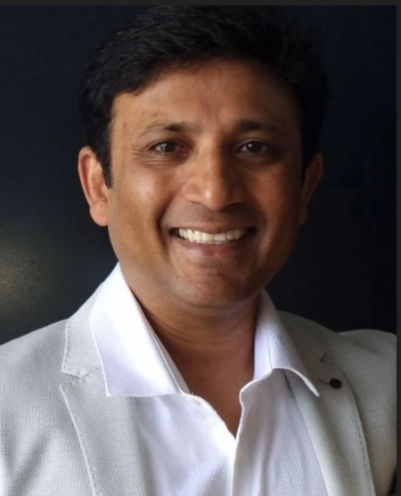 A smiling man with short black hair in a white blazer and shirt against a dark background.