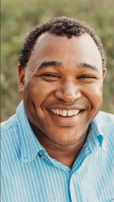 Close-up of a smiling man with short curly hair wearing a light blue striped shirt, outdoors with blurry green background.