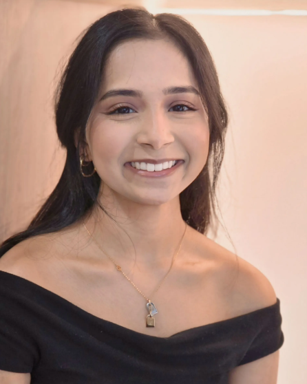 A smiling woman with long dark hair, wearing a black off-shoulder top, gold hoop earrings, and a gold necklace with a padlock pendant, standing against a plain beige background.