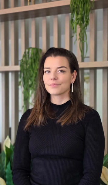 Young woman with brown hair and earrings, standing indoors with a background of vertical wooden slats and hanging green plants.