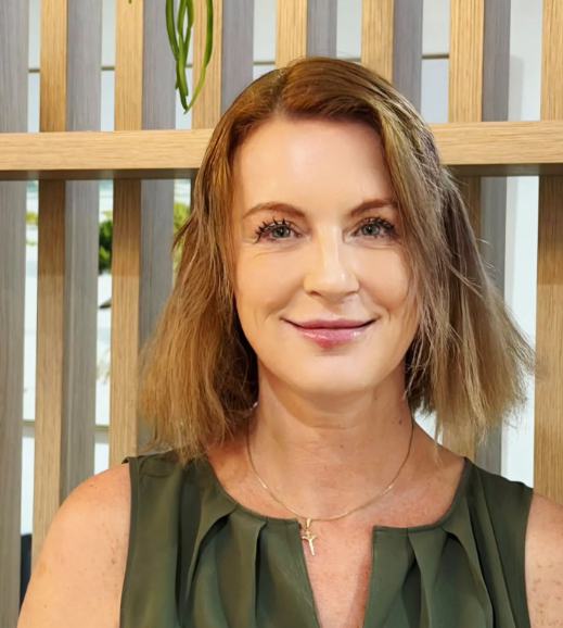 A woman with shoulder-length light brown hair smiling, sitting in front of a wooden slat wall with some green plants visible.