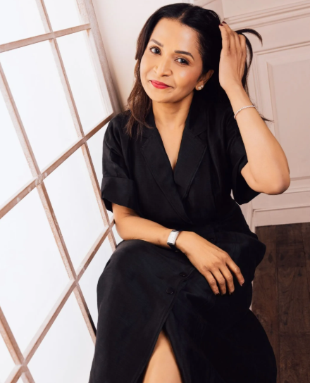 A woman with shoulder-length dark hair sitting near a window, wearing a black dress, smiling and touching her hair.