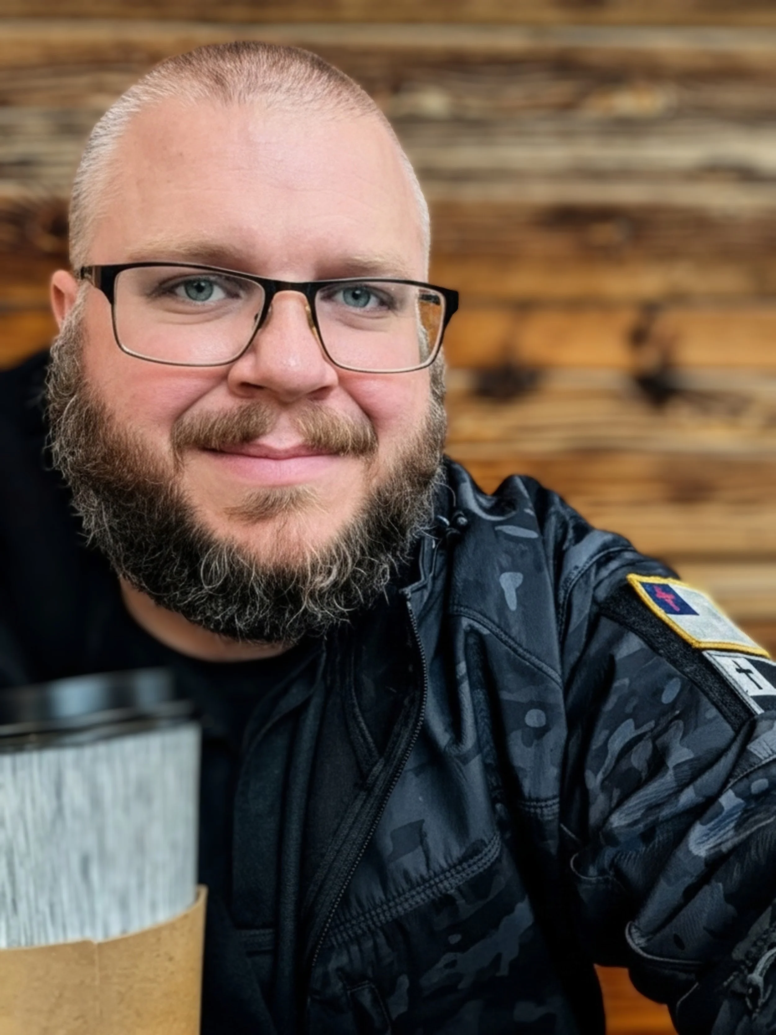 A man with glasses and a beard, wearing a police uniform, smiling, sitting in front of a wooden wall, with a coffee cup in the foreground.