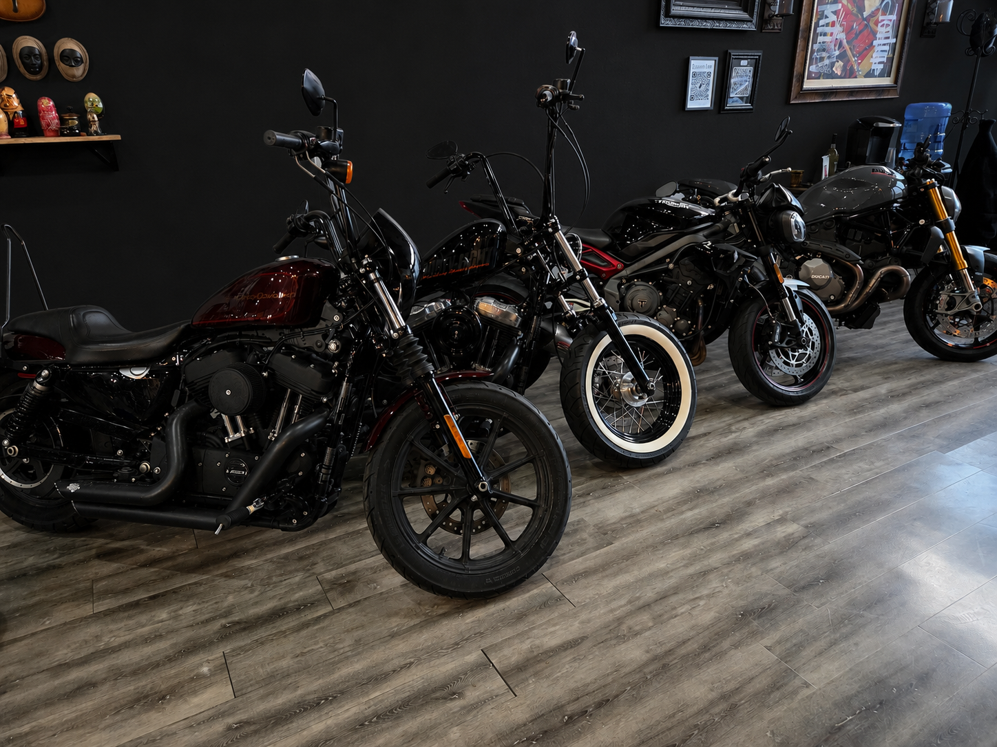 Three motorcycles parked indoors on a wooden floor, with a dark wall behind them decorated with framed pictures and a shelf holding figurines. Harley Sportster, Triumph, Ducati