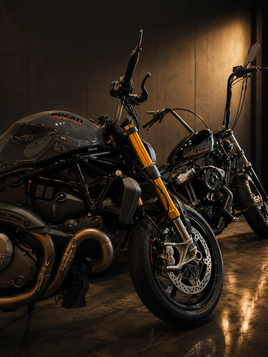 Harley Sportster and Ducati Monster motorcycles parked indoors with a dark background and polished concrete floor.