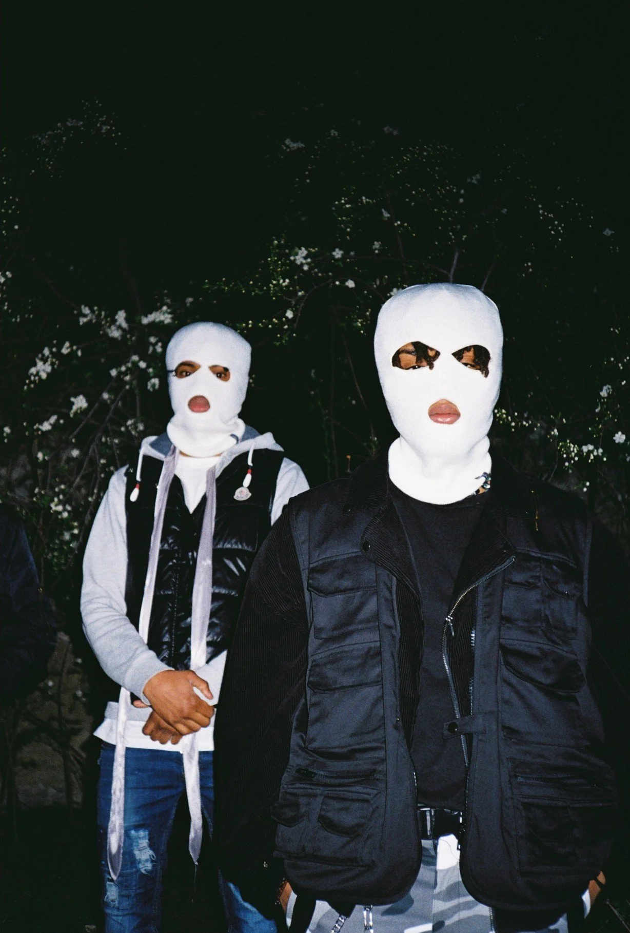Two people wearing white ski masks in front of a dark leafy background, one with a black vest and the other with a gray hoodie and backpack.
