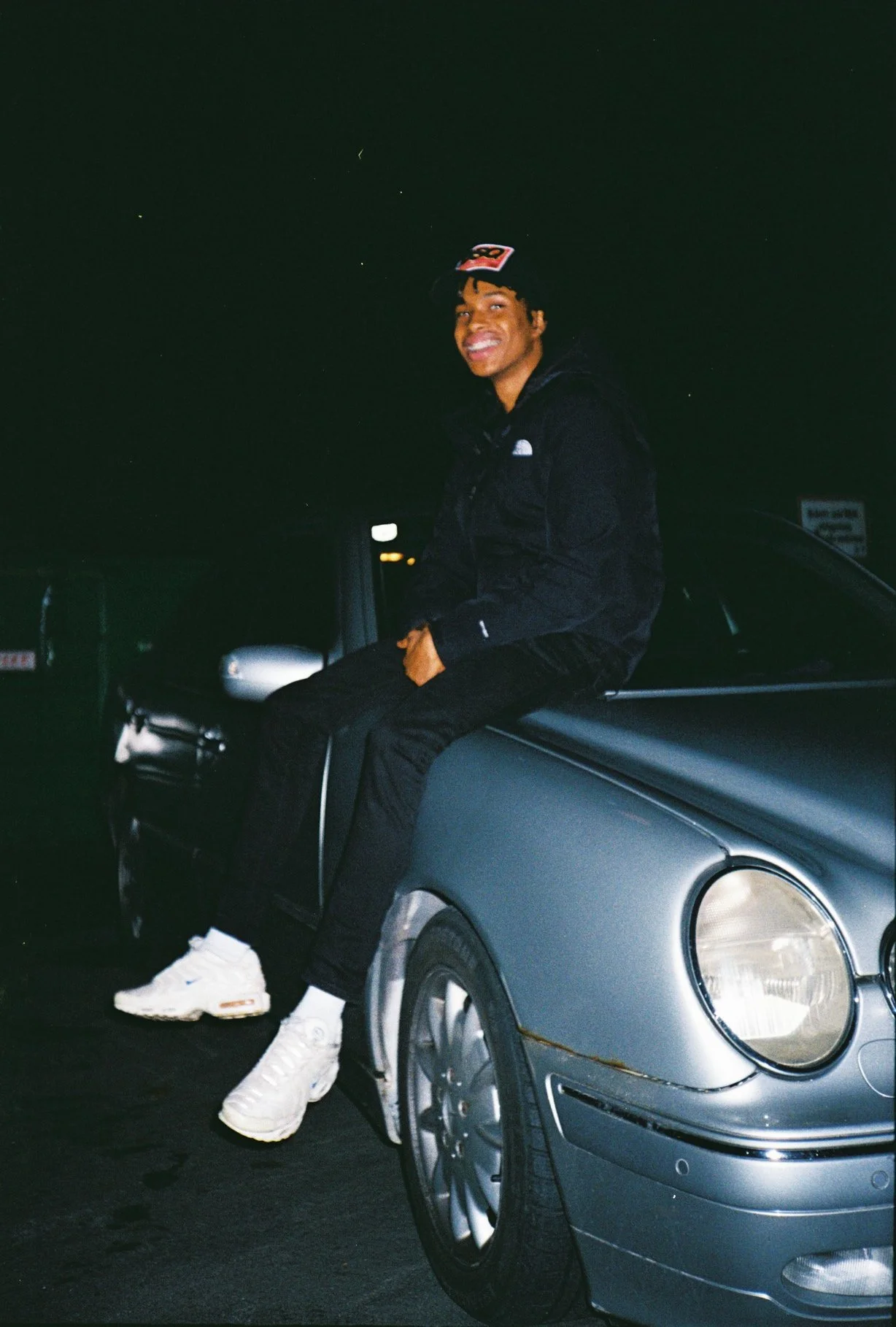 A young man sitting on the hood of a silver car at night, smiling at the camera. He is wearing a black hoodie, black pants, white sneakers, and a black cap.