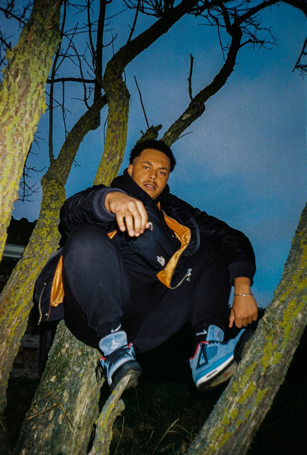 A person sitting in a tree at dusk, wearing a black jacket and sneakers, with a serious expression, surrounded by branches and a blue sky.
