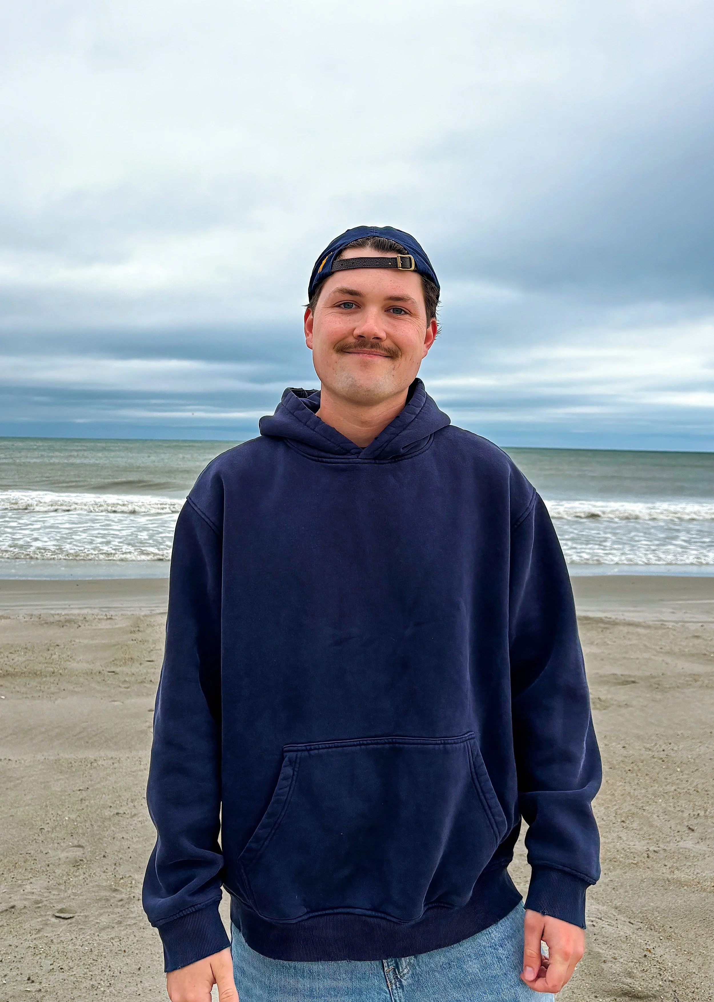A young man with light skin, dark hair, and a mustache wearing a navy blue hoodie and a backward navy baseball cap standing on a beach with the ocean and cloudy sky in the background.