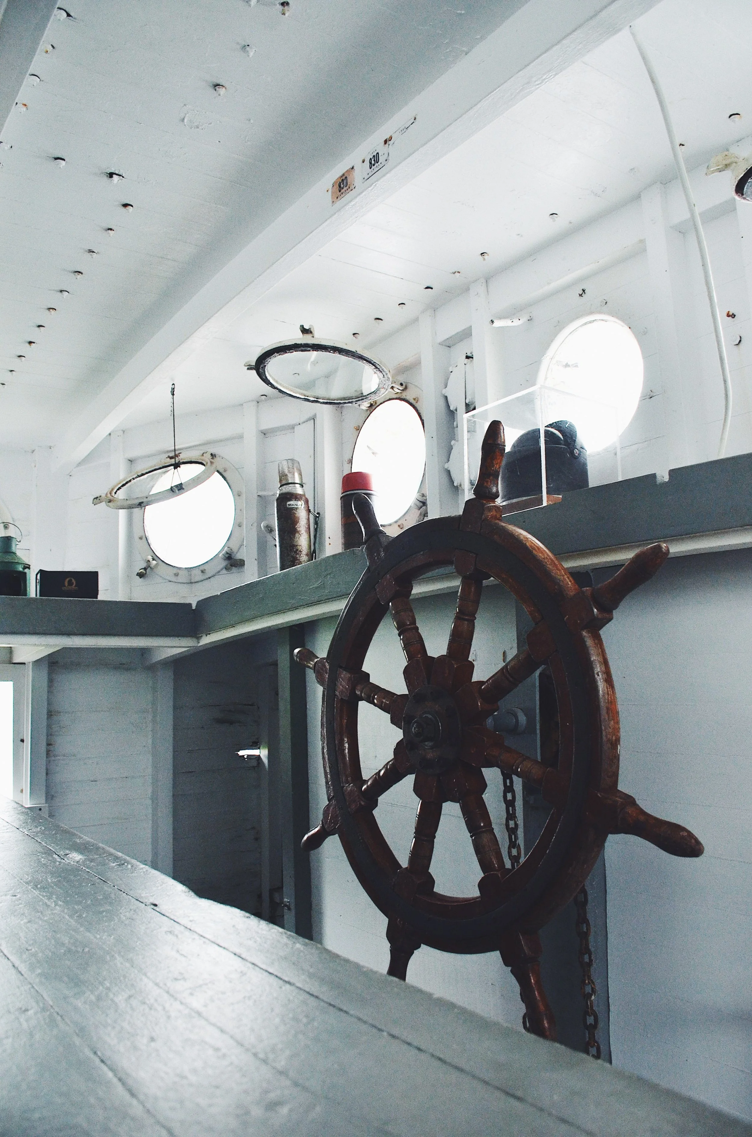 Interior of a boat with a wooden ship wheel, circular portholes letting in light, and various objects on a shelf.