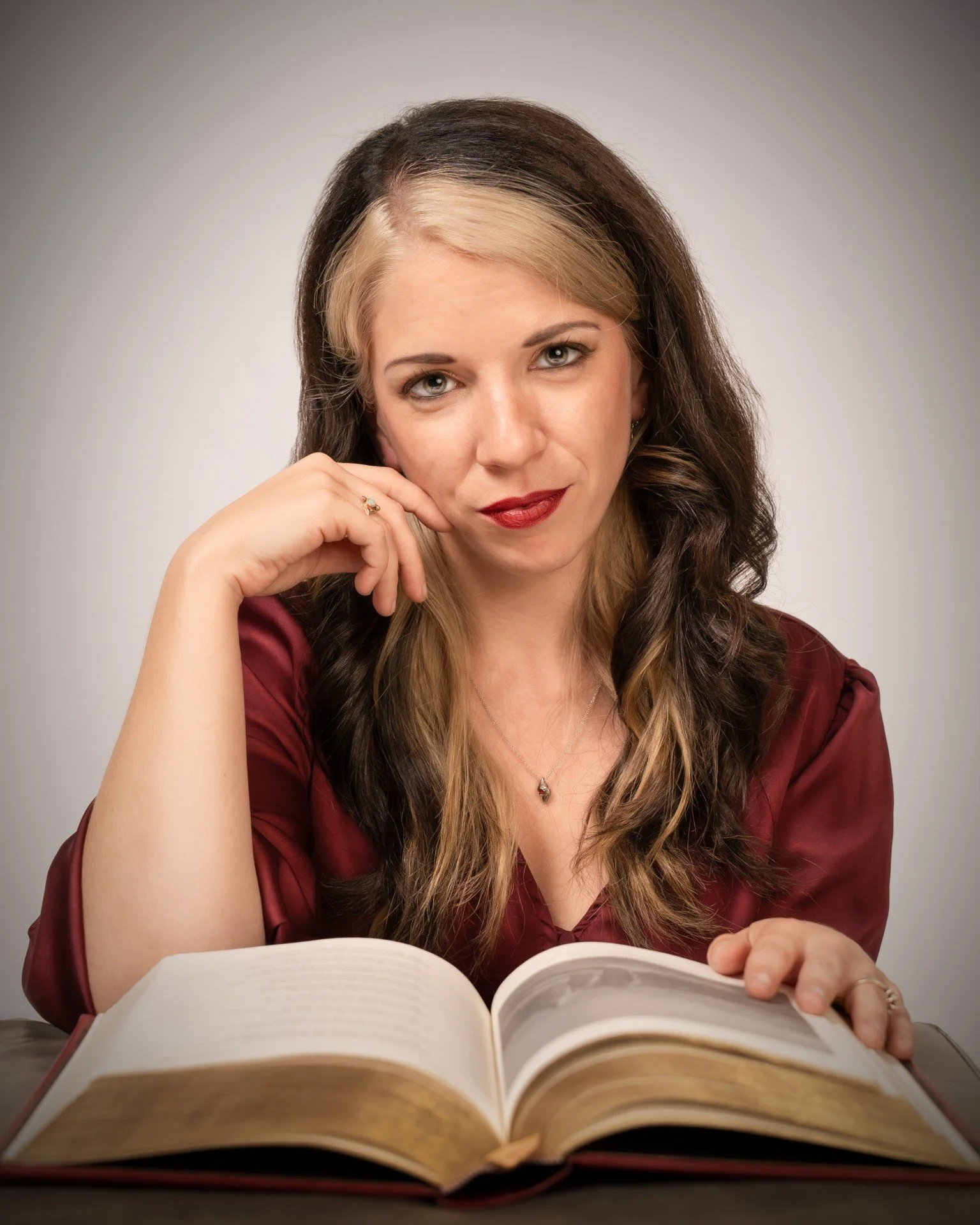 A woman with long, wavy brown hair with blonde highlights reading a large open book.