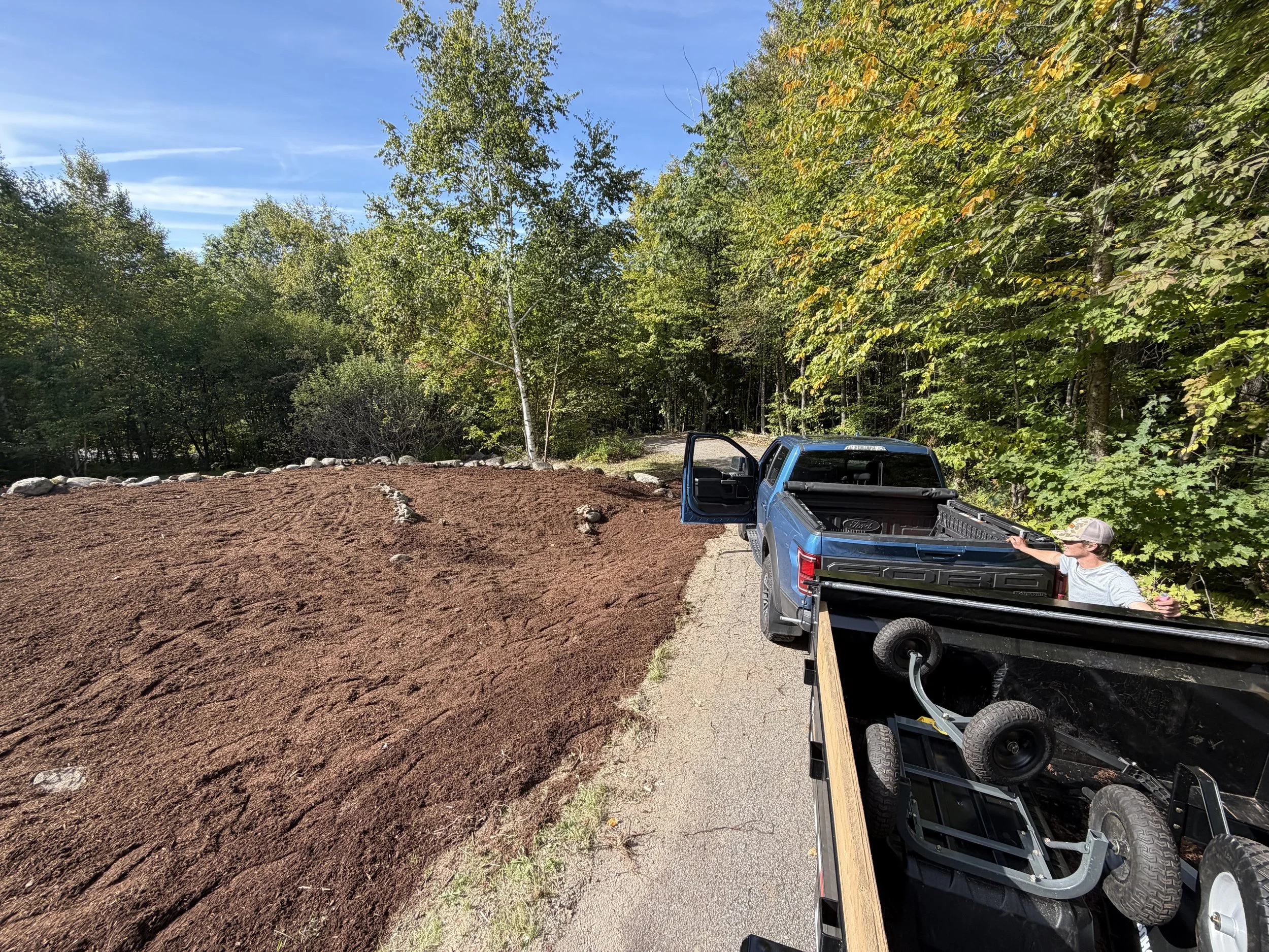 A dirt area with a border of rocks, a parked blue pickup truck with its tailgate down, a person wearing a cap and sunglasses near the truck, and a forested background with green trees under a clear blue sky.