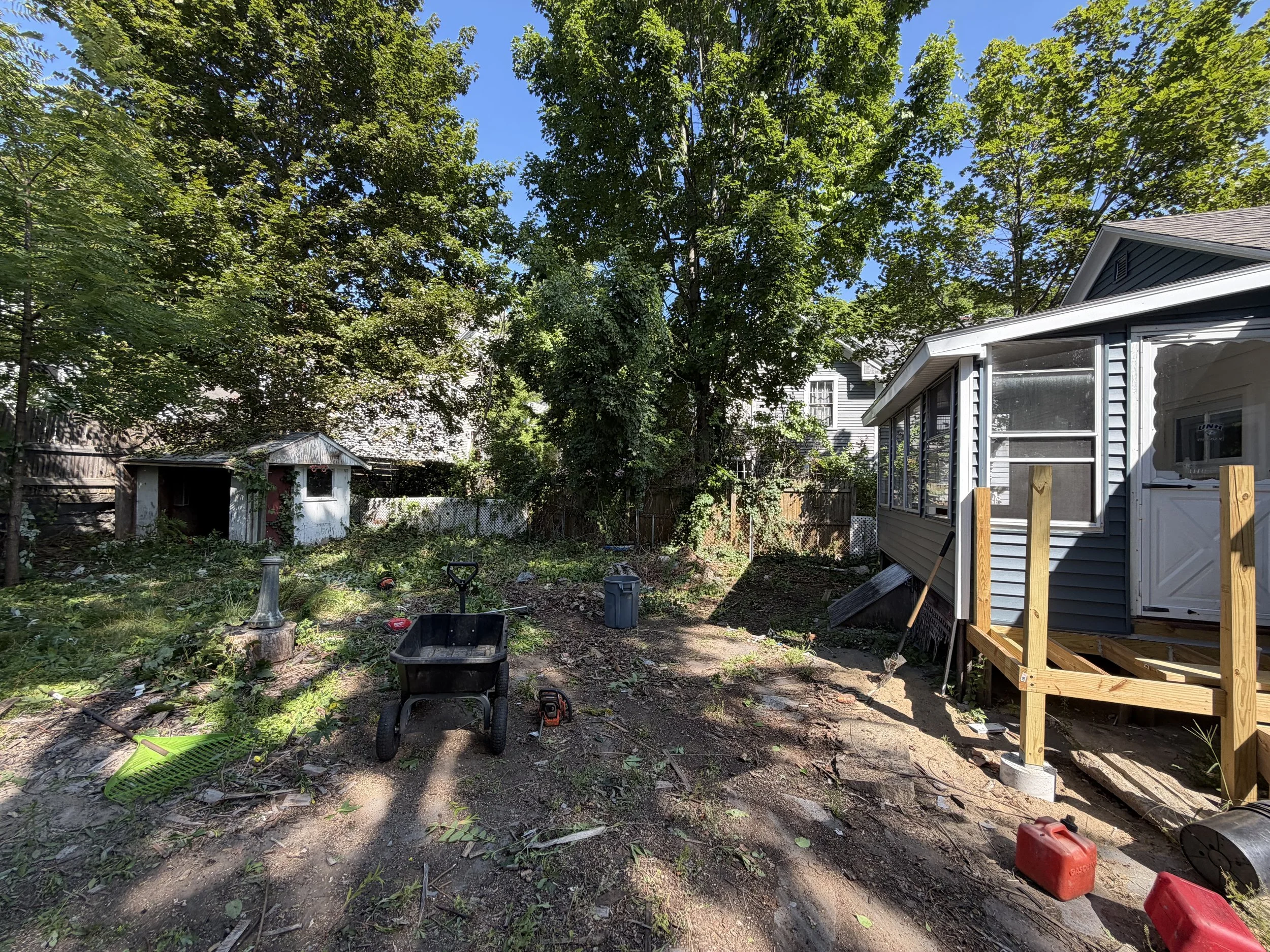 A backyard yard with trees, a shed, and a partially constructed wooden deck. Gardening tools and other objects are scattered around the yard.