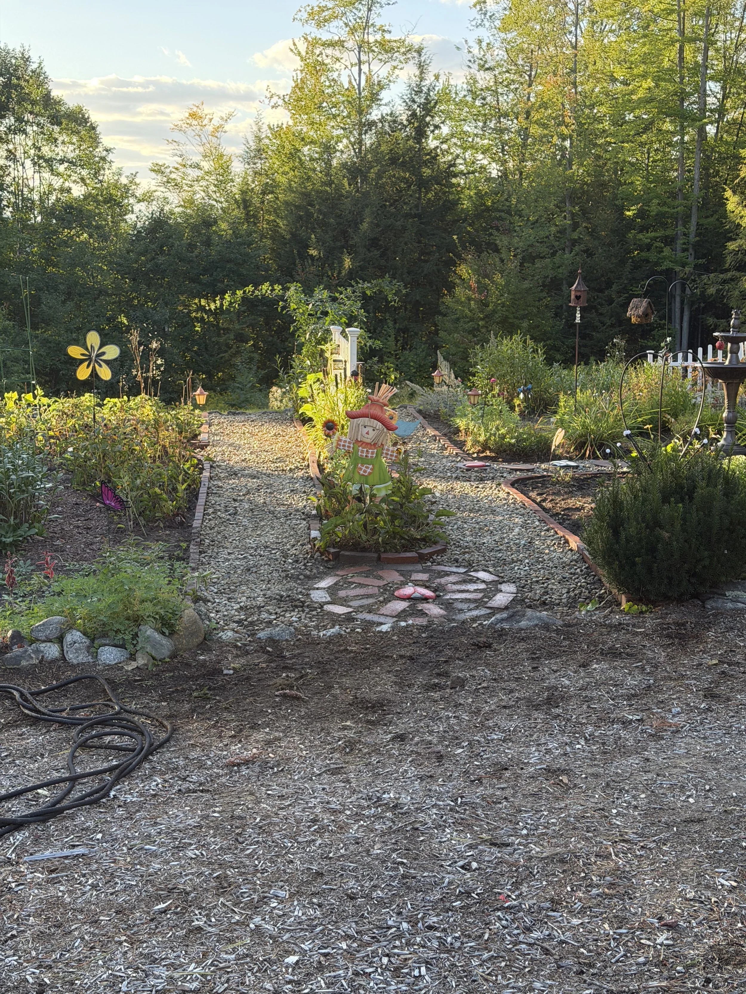 A garden with a dirt and mulch pathway leading to a decorative scarecrow statue, surrounded by plants and flowers, with trees and a blue sky with clouds in the background.