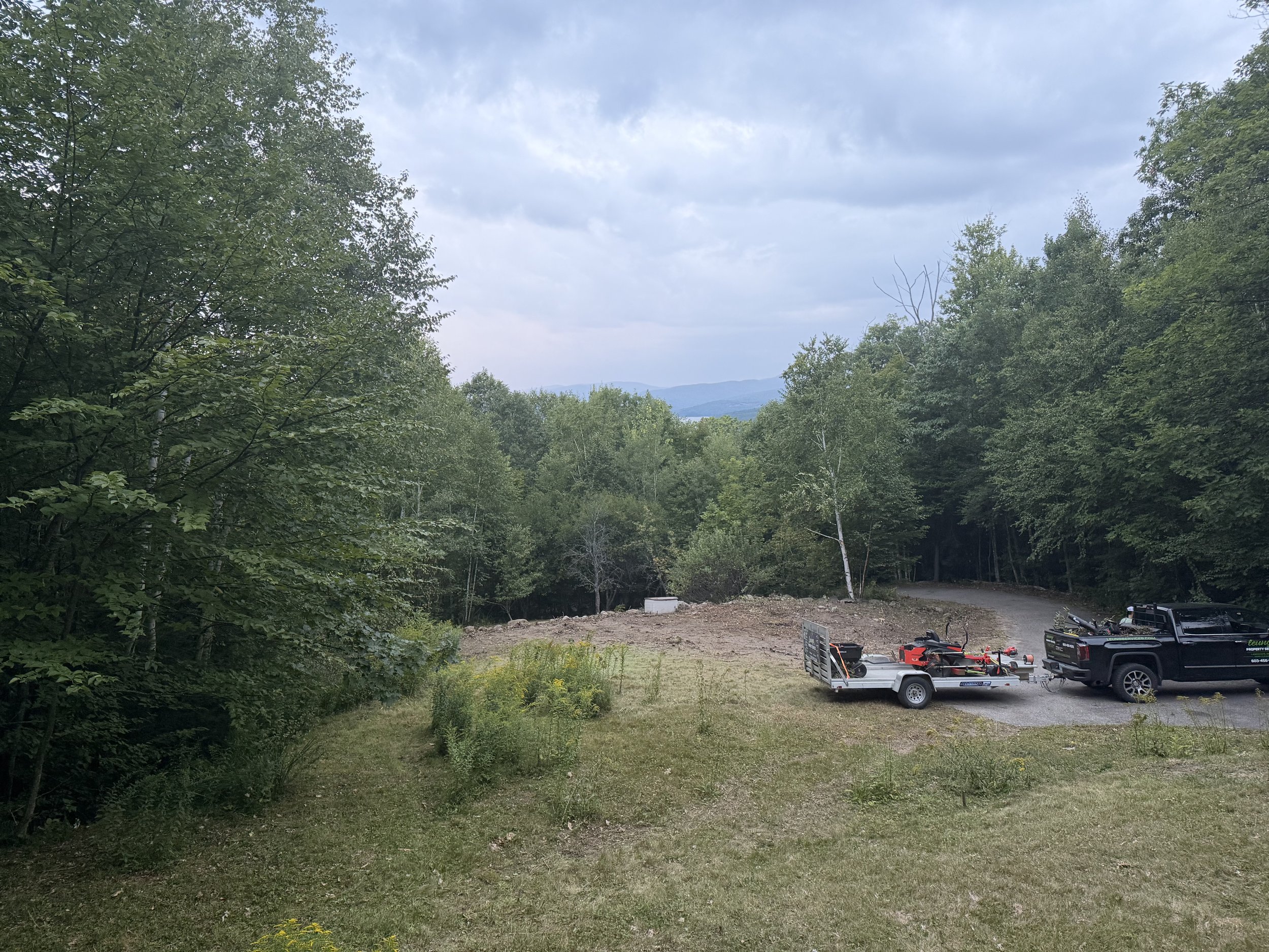 A mountainous landscape with trees and clouds in the sky, a flat dirt area, and parked vehicles and trailers at the bottom right corner.
