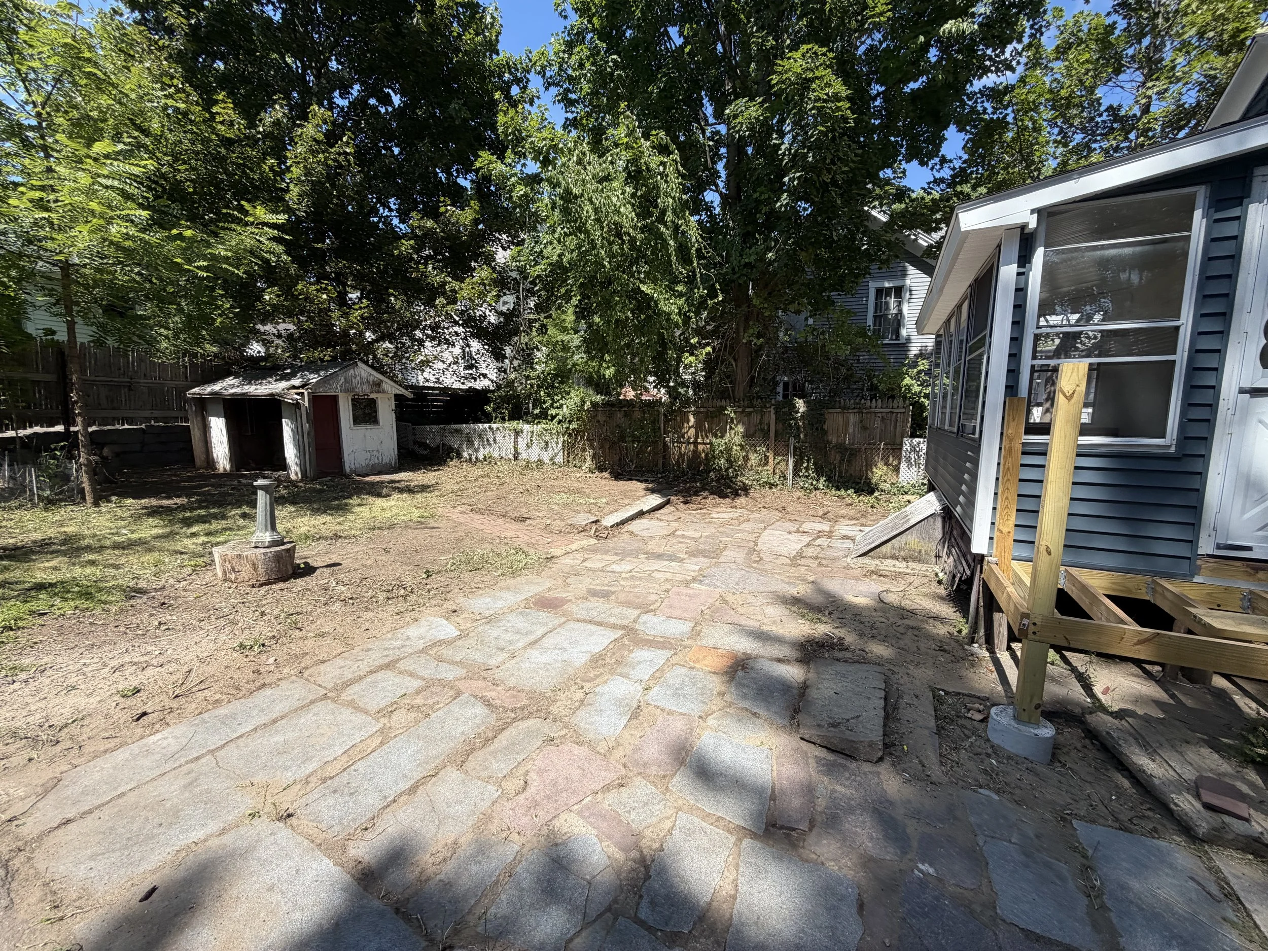 A backyard area features a large tree providing shade, a small white shed with a red door, a raised deck with stairs, a patch of dirt, and a stone pathway. There's a wooden fence surrounding the yard and residential houses visible in the background.
