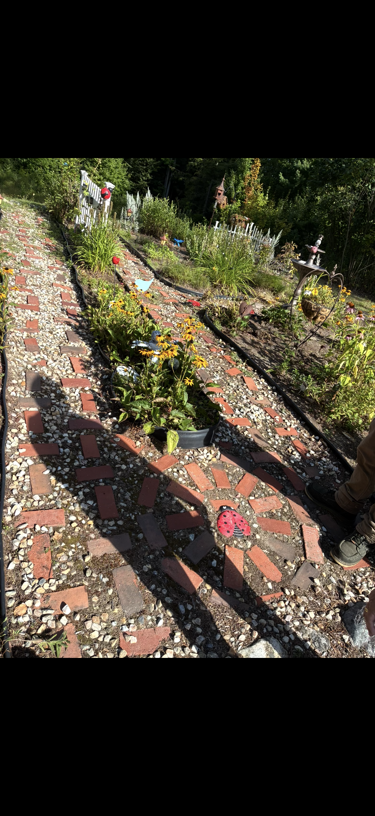 A garden with flower beds, a brick and gravel pathway, decorative girl-themed ladybug garden ornaments, a white picket fence, and a small playhouse in the background, all surrounded by green trees.