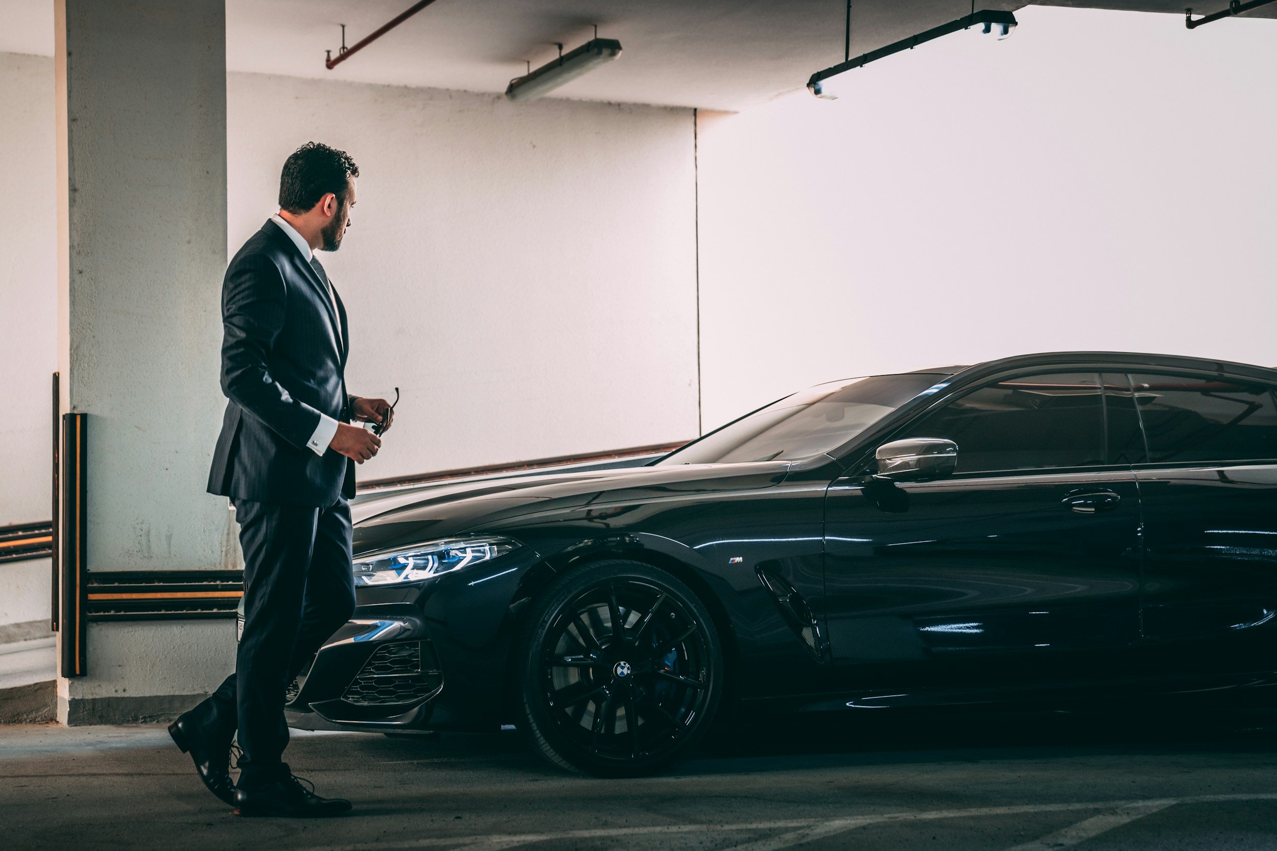 A man dressed in a suit stands beside a black luxury car in a parking garage.