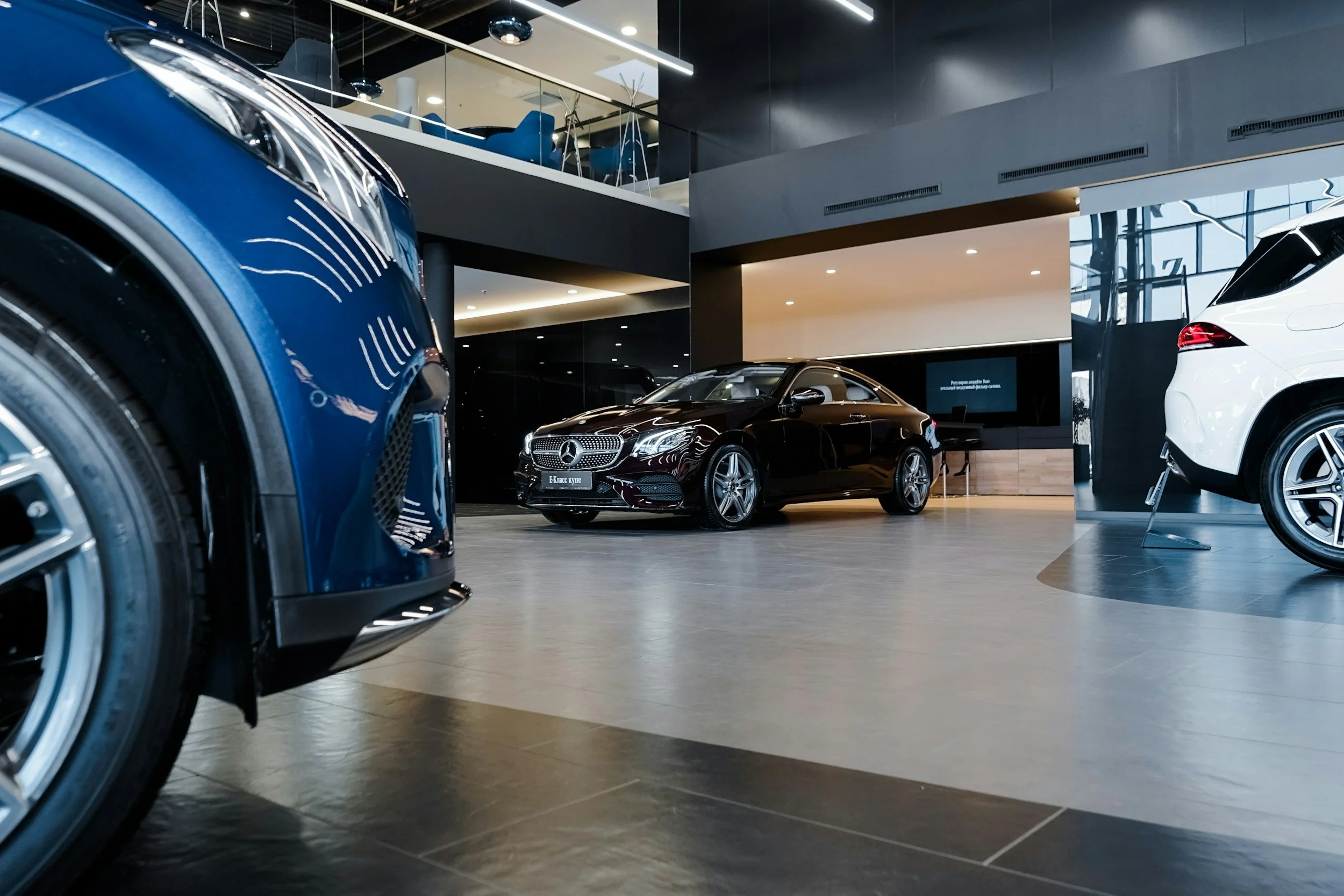 Interior of a modern car dealership showing a red luxury Mercedes-Benz car in the center, a white SUV on the right, and parts of a blue vehicle on the left, with a seating area upstairs visible through glass.