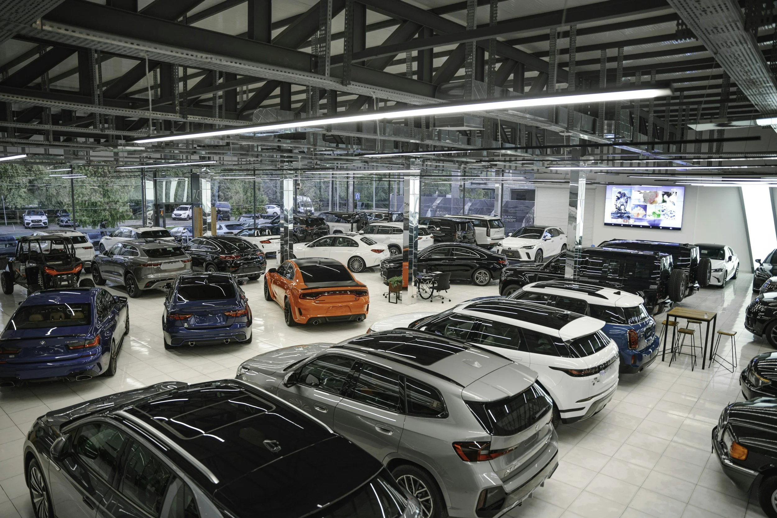 Interior of a modern car dealership showroom filled with various new cars, with large glass windows letting in natural light and digital screens on the walls.