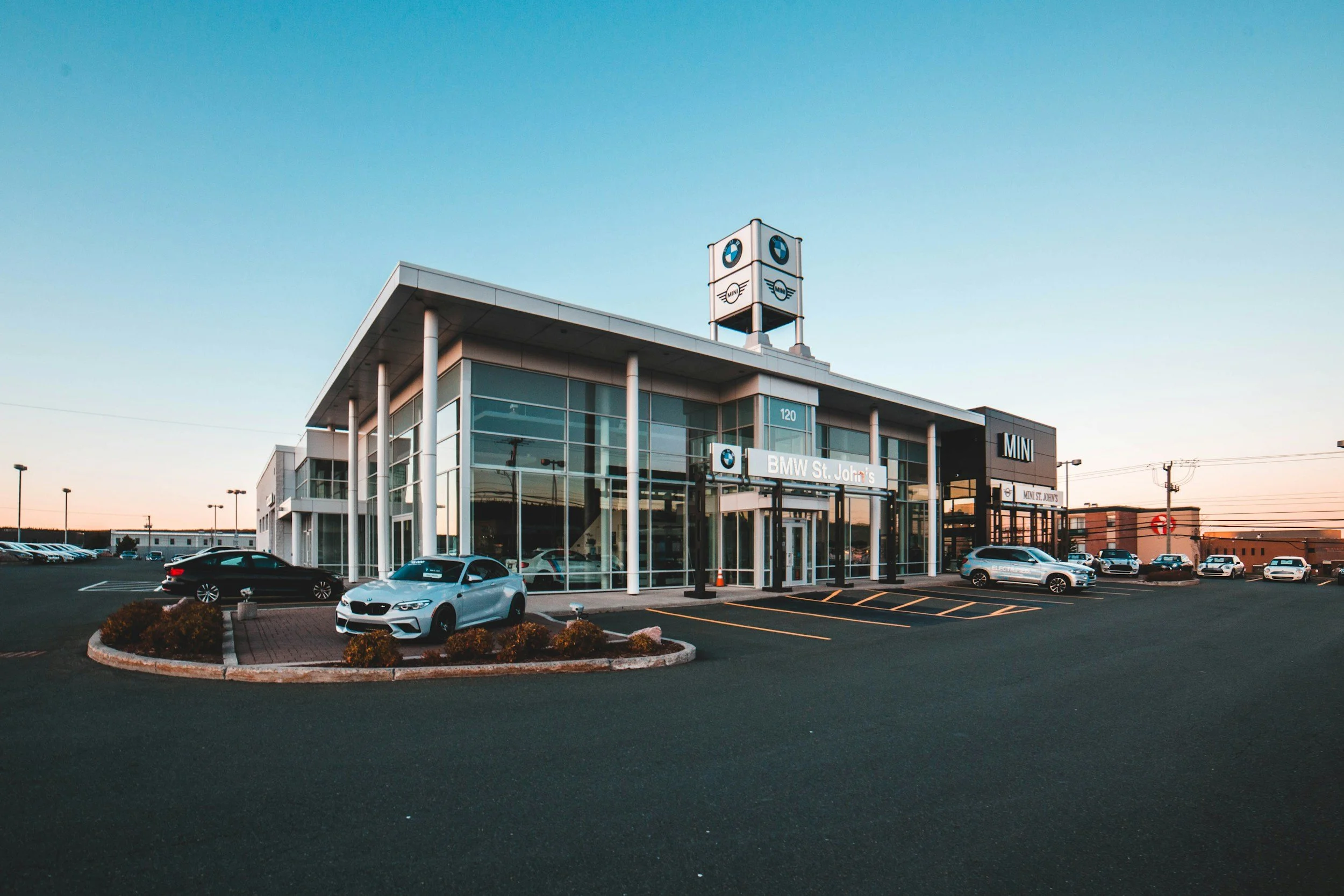 Modern car dealership with a BMW sign and several new vehicles parked outside, under a clear blue sky at sunset.