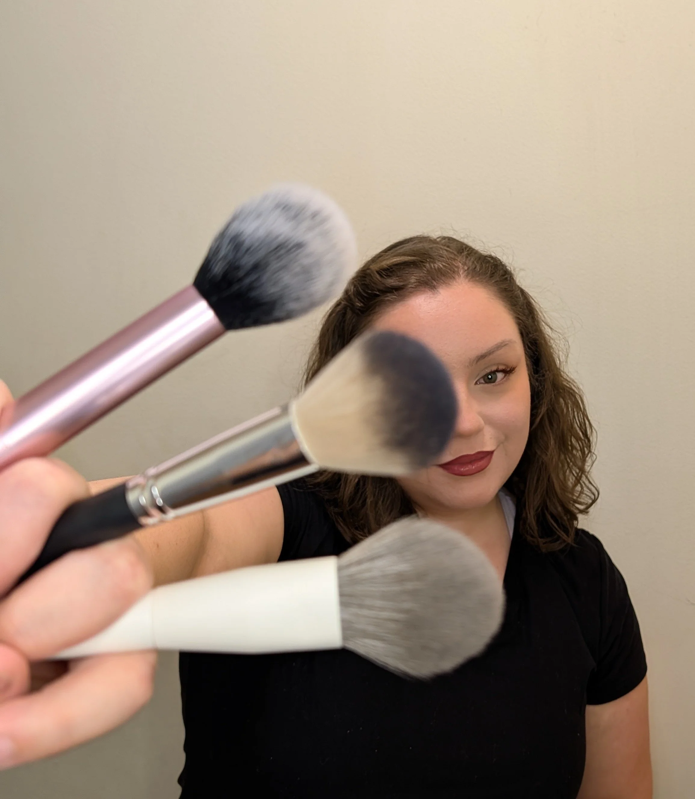 Close-up of a woman with brown curly hair applying makeup, holding makeup brushes, and looking at the camera against a beige wall.