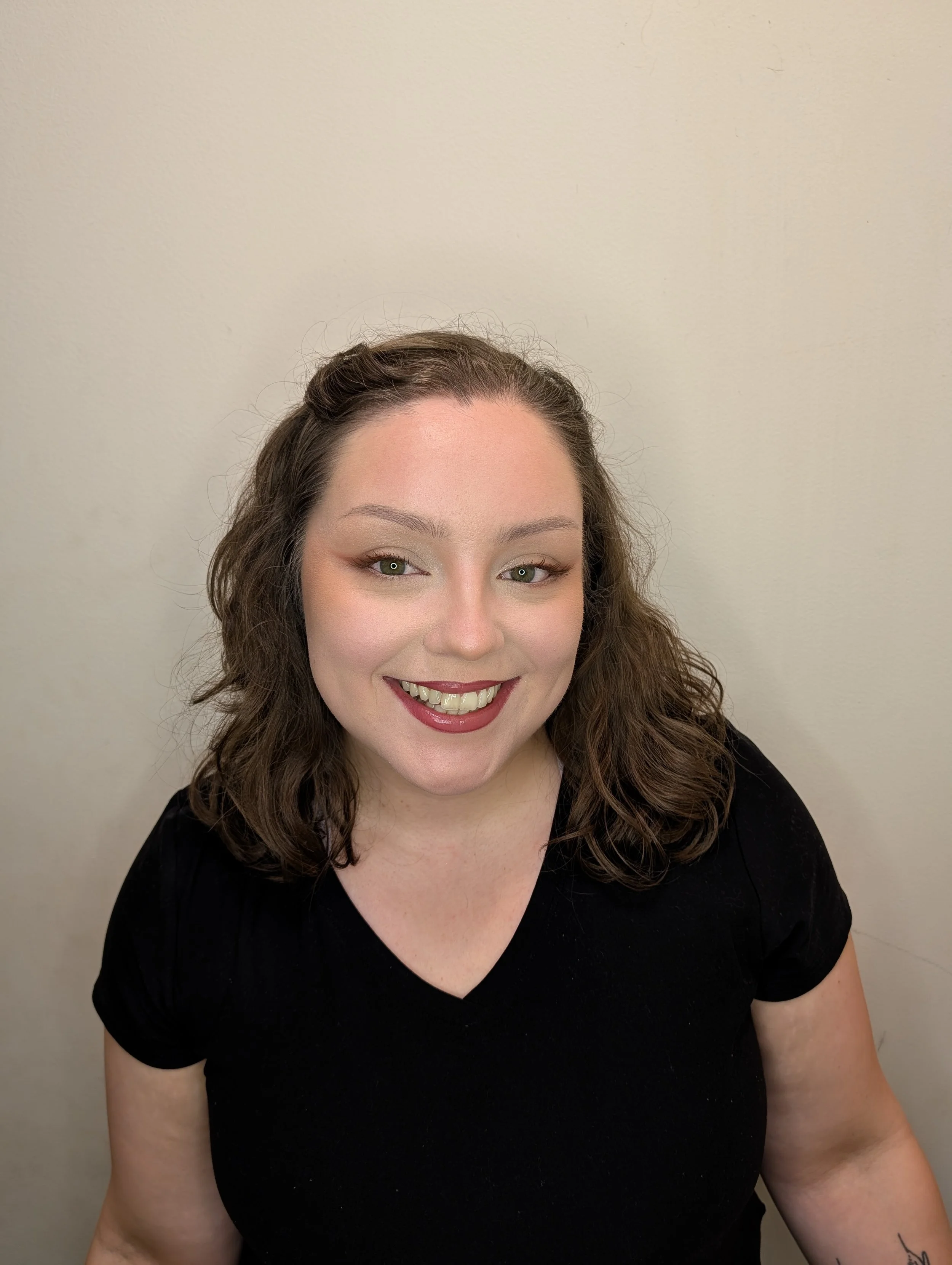 Close-up photo of a smiling woman with wavy brown hair and fair skin, wearing a black top, against a plain light-colored wall.