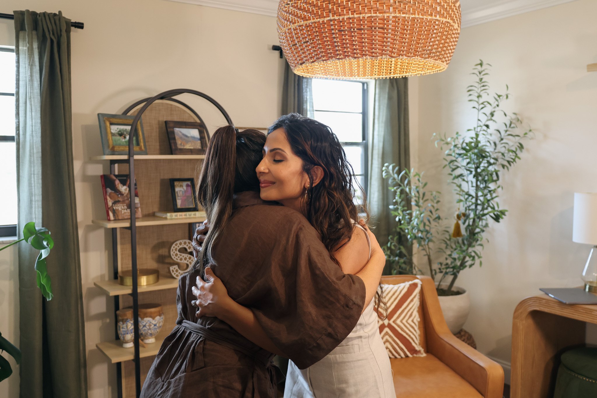 Two women hugging in a living room, with a bookshelf, houseplants, and a window with curtains in the background.