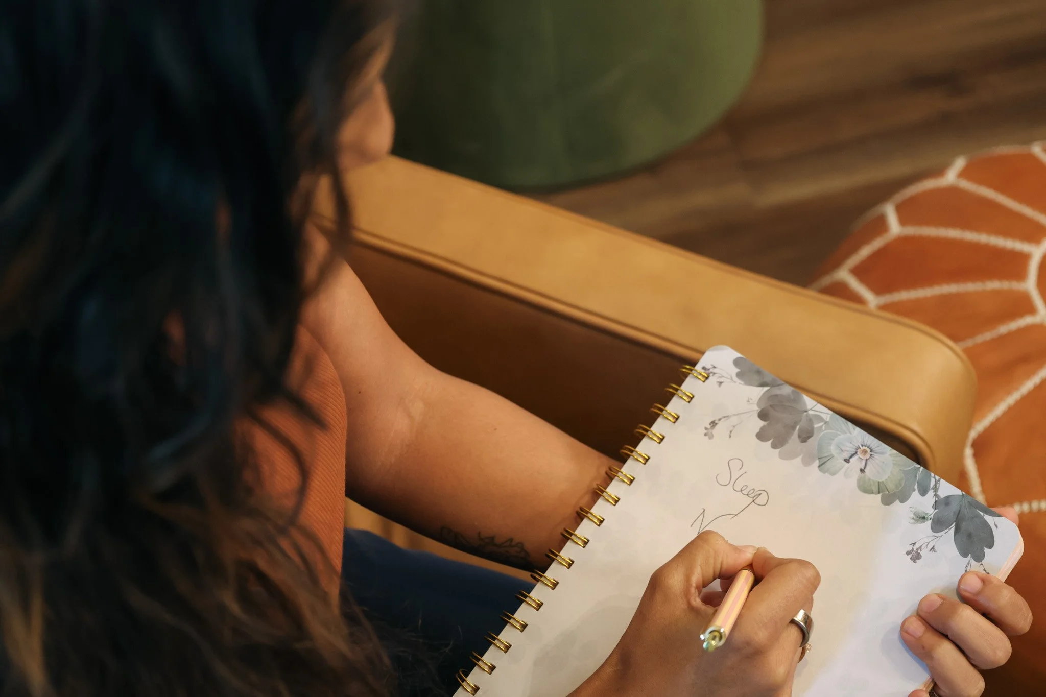 Person with long dark hair writing in a spiral-bound notebook with floral cover, holding a pink pen, sitting at a wooden table.