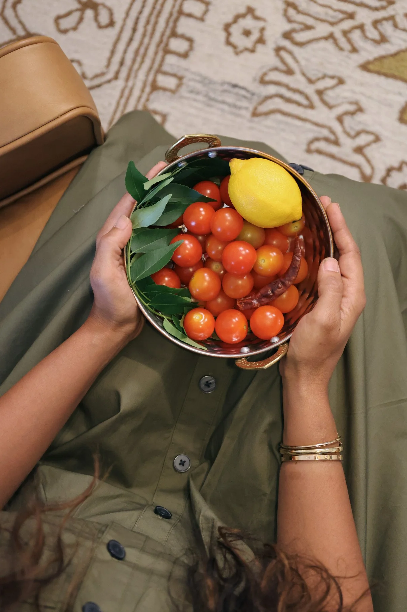 Hand holding a small basket of cherry tomatoes, a lemon, a dried chili, and green leaves, with a person sitting on a patterned rug.
