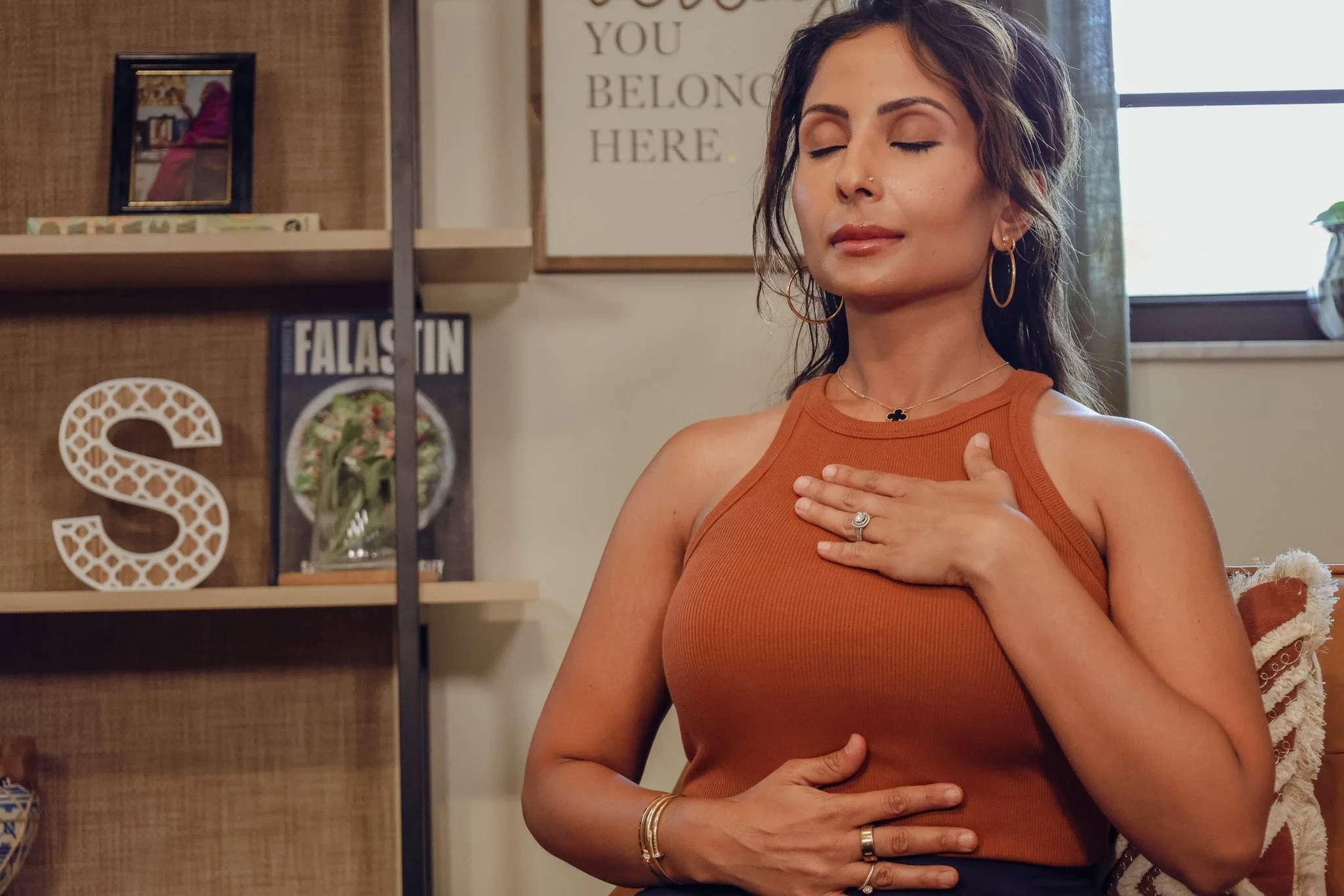 Woman with long dark hair and gold jewelry, eyes closed, hand on chest, wearing an orange sleeveless top, indoors with a bookshelf and decorative items in the background.